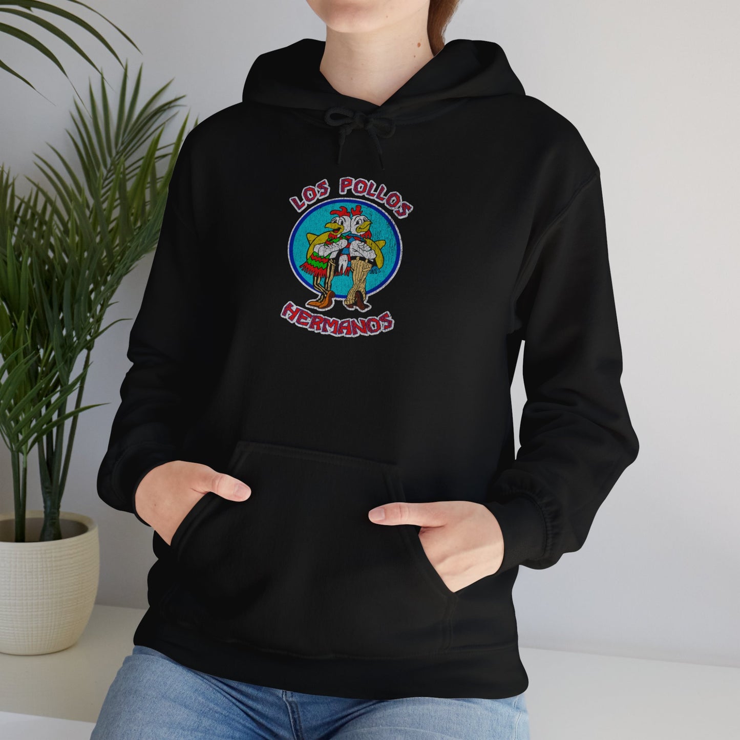 Female model wearing a black Soft Unisex Hoodie Heavy Blend Hooded Sweatshirt featuring the Los Pollos Hermanos logo from the TV show Breaking Bad and Better Call Saul in a vintage distressed style. Logo is printed on both front and back of hoodie.