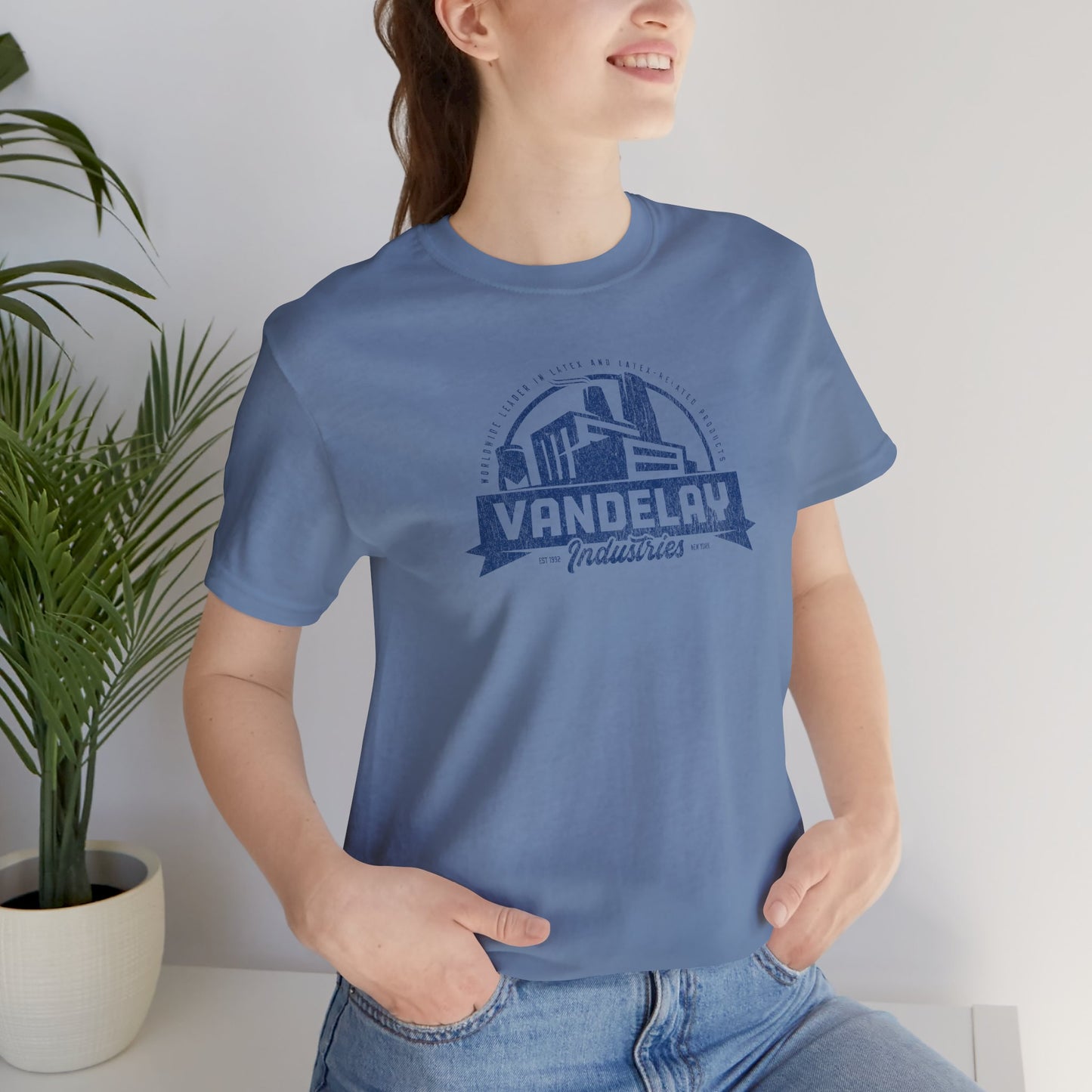 Female model wearing a steel blue Soft Bella+Canvas 3001 short-sleeve t-shirt tee featuring a Vandelay Industries logo graphic, uniquely designed by ScriptedBrand in a vintage distressed style. The fictional company is part of the George Costanza storyline in the TV show Seinfeld.
