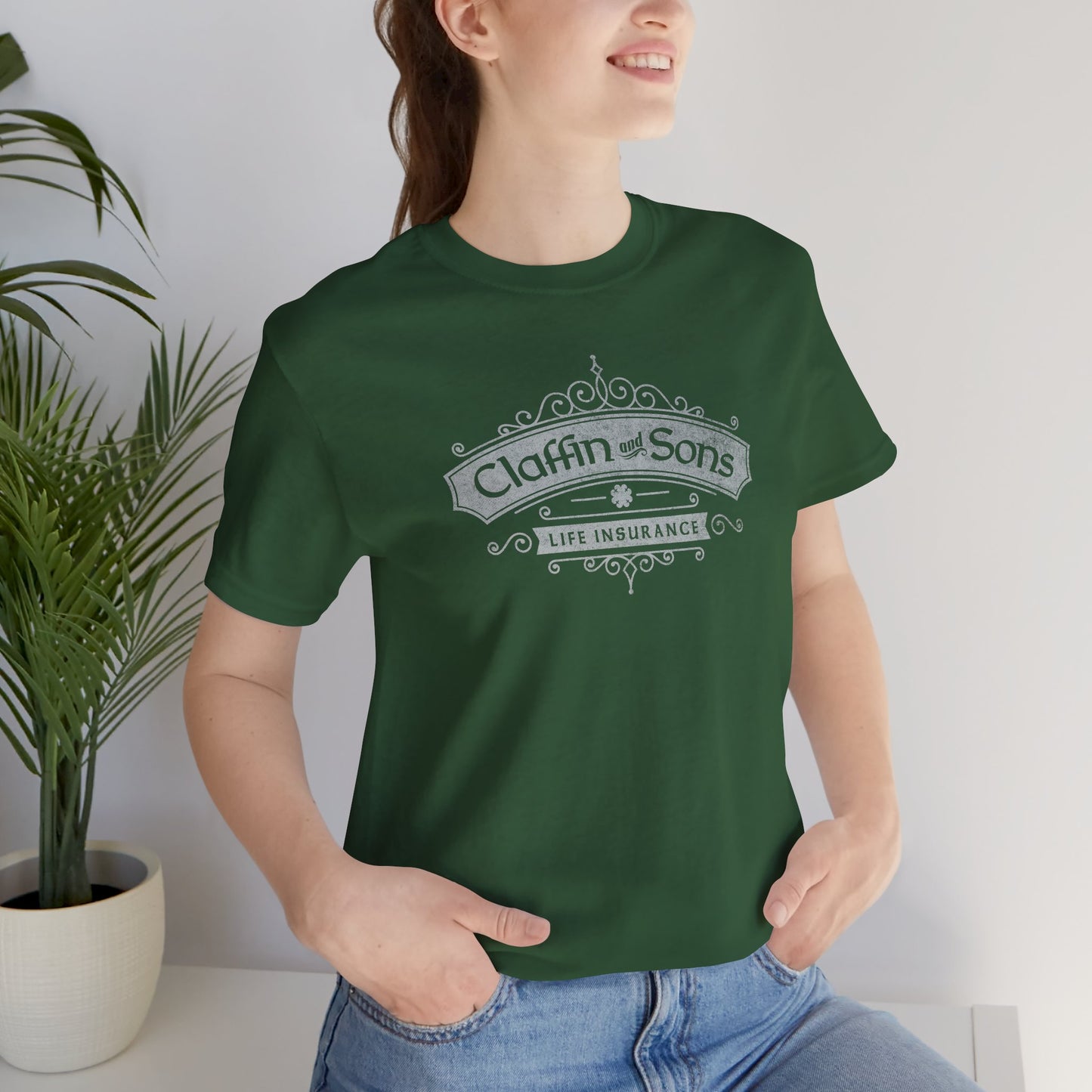 Female model wearing an evergreen colored soft Bella+Canvas 3001 short-sleeve t-shirt featuring a logo graphic of Claffin & Sons Insurance, designed by ScriptedBrand in a vintage distressed vintage distressed style, inspired by the TV show Bad Sisters.