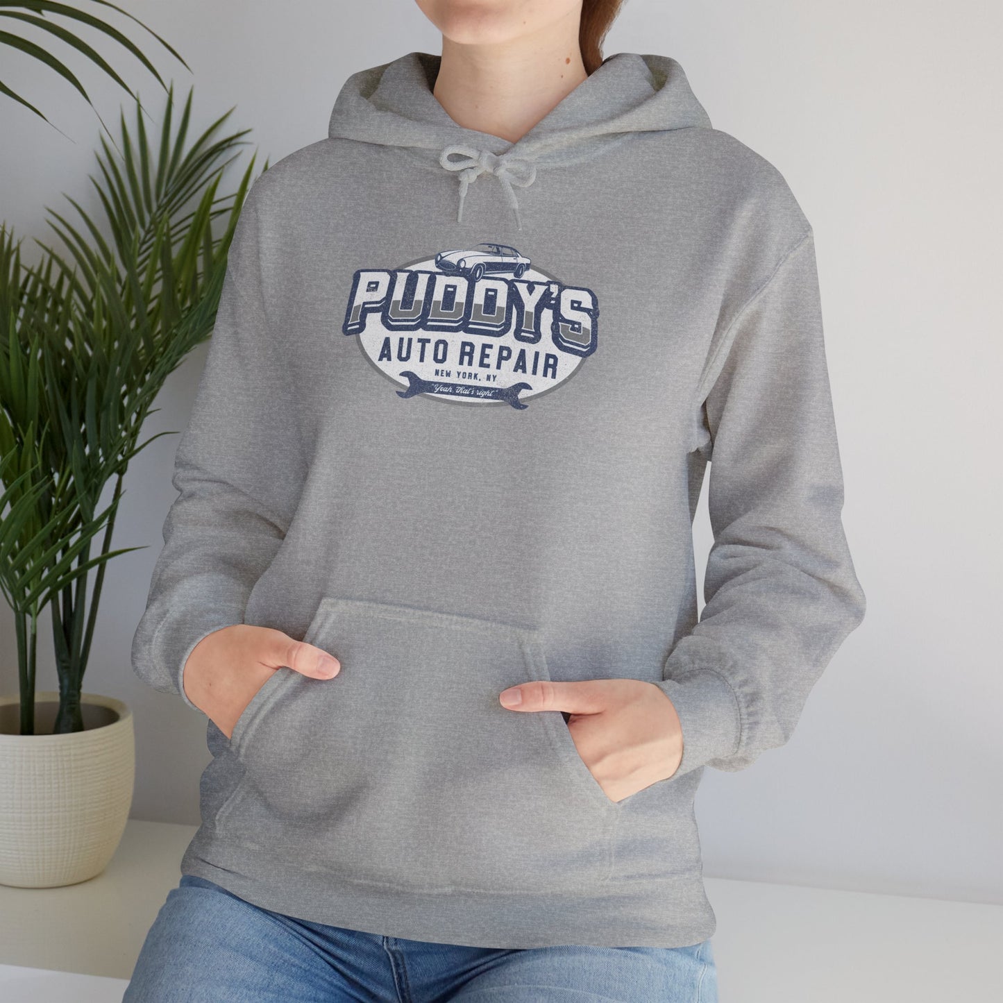 Female model wearing a sport heather gray soft Unisex Hoodie Heavy Blend Hooded Sweatshirt featuring a Puddy's Auto Repair graphic designed by ScriptedBrand inspired by the TV show Seinfeld in a vintage distressed style.