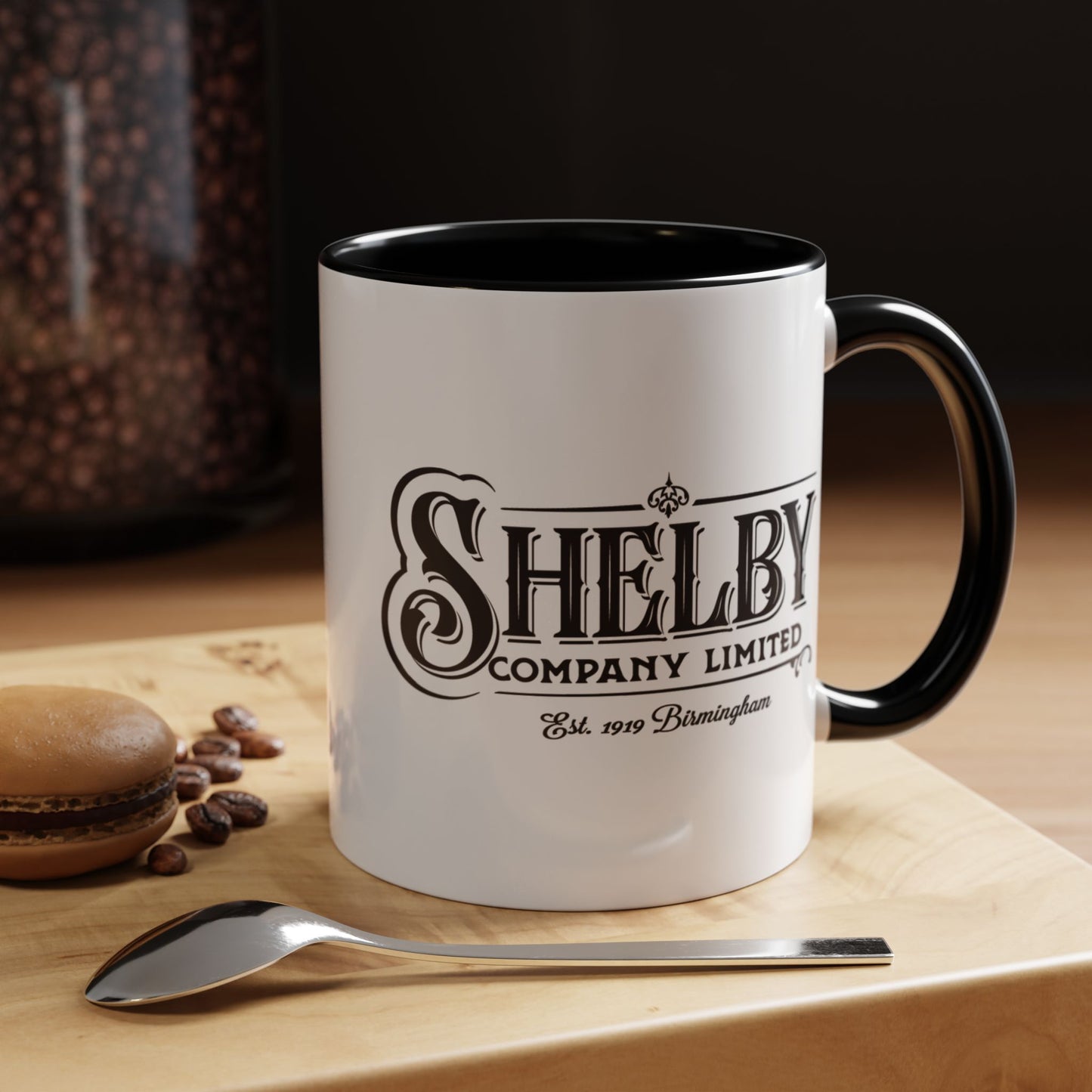 Front view of a 11 oz White Ceramic Mug with black handle/interior featuring the fictional Shelby Company Limited Logo from the TV show Peaky Blinders printed on both sides. Graphic is designed by ScriptedBrand and includes the text "Est. 1919 Birmingham". Mug is shown placed in a kitchen setting.
