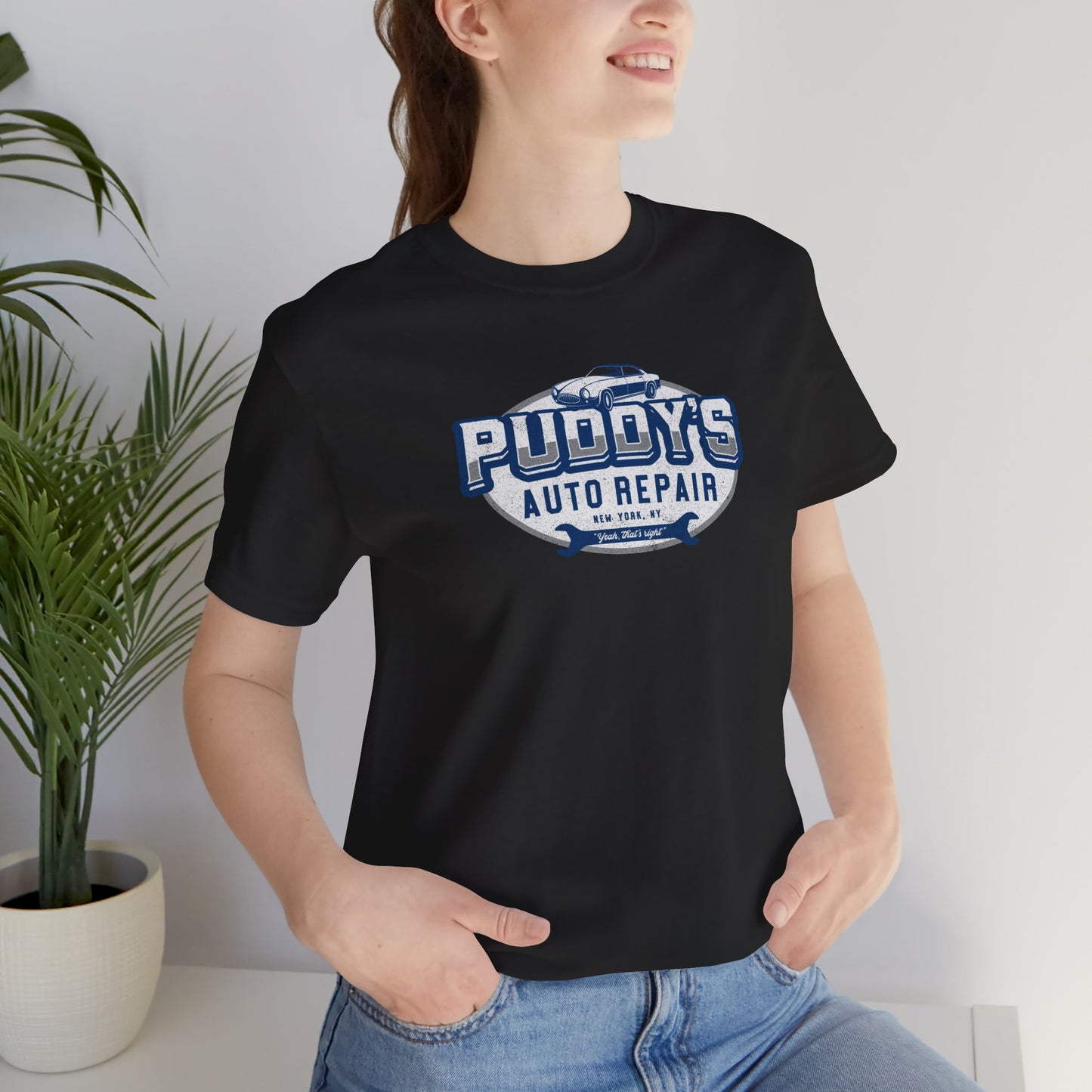 Female model wearing a black soft short-sleeve t-shirt featuring a Puddy's Auto Repair graphic designed by ScriptedBrand inspired by the TV show Seinfeld in a vintage distressed ink-stamped worn-in style.