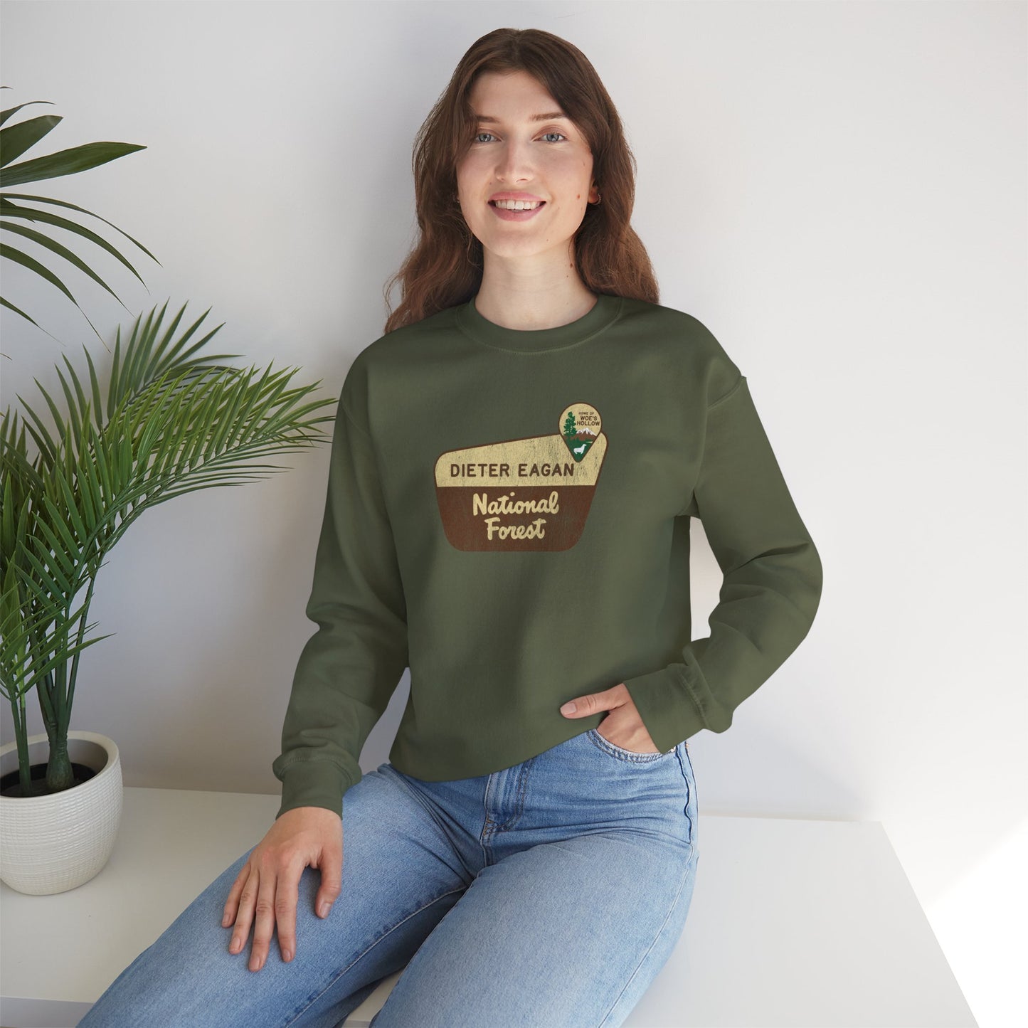 Female model wearing a military green Soft Unisex Gildan 18000 sweatshirt featuring a logo graphic of the Dieter Eagan National Forest sign designed by ScriptedBrand, inspired by the TV show Severance in a vintage distressed style.