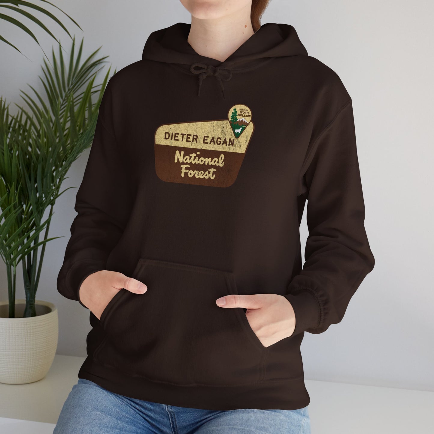 Female model wearing a dark chocolate brown colored Soft Unisex Hoodie Heavy Blend Hooded Sweatshirt featuring the logo graphic of the Dieter Eagan National Forest inspired by the TV show Severance in a vintage distressed ink-stamped worn-in style.