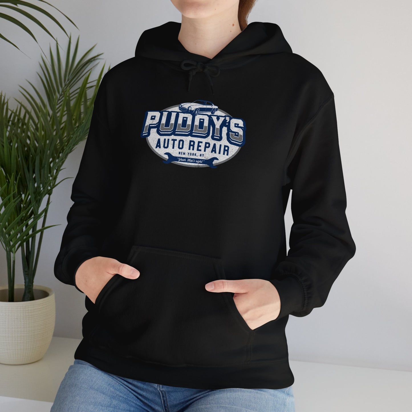 Female model wearing a black soft Unisex Hoodie Heavy Blend Hooded Sweatshirt featuring a Puddy's Auto Repair graphic designed by ScriptedBrand inspired by the TV show Seinfeld in a vintage distressed style.
