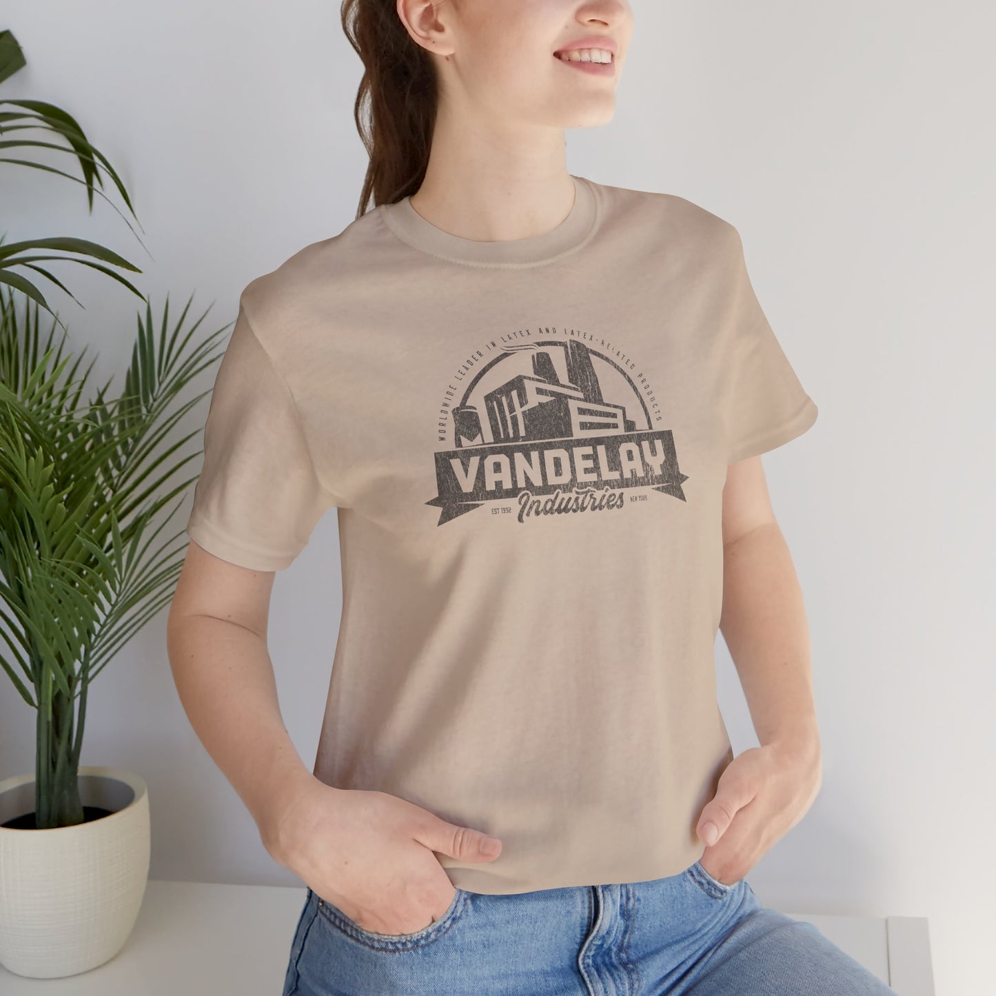 Female model wearing a tan Soft Bella+Canvas 3001 short-sleeve t-shirt tee featuring a Vandelay Industries logo graphic, uniquely designed by ScriptedBrand in a vintage distressed style. The fictional company is part of the George Costanza storyline in the TV show Seinfeld.