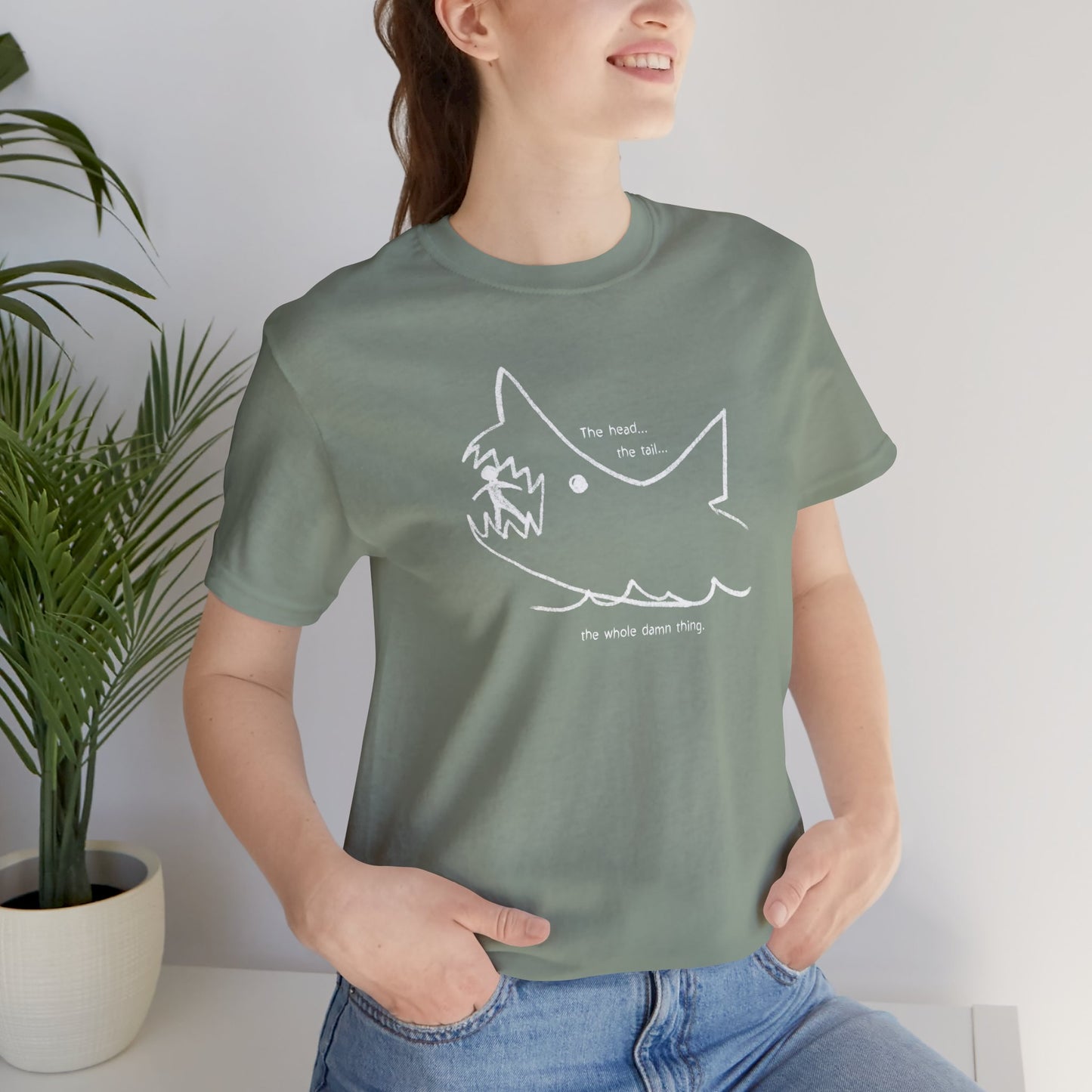 Female model wearing a sage Green colored soft short-sleeve Bella+Canvas 3001 t-shirt featuring the chalkboard drawing of a shark eating a person, drawn by Quint in the movie Jaws.