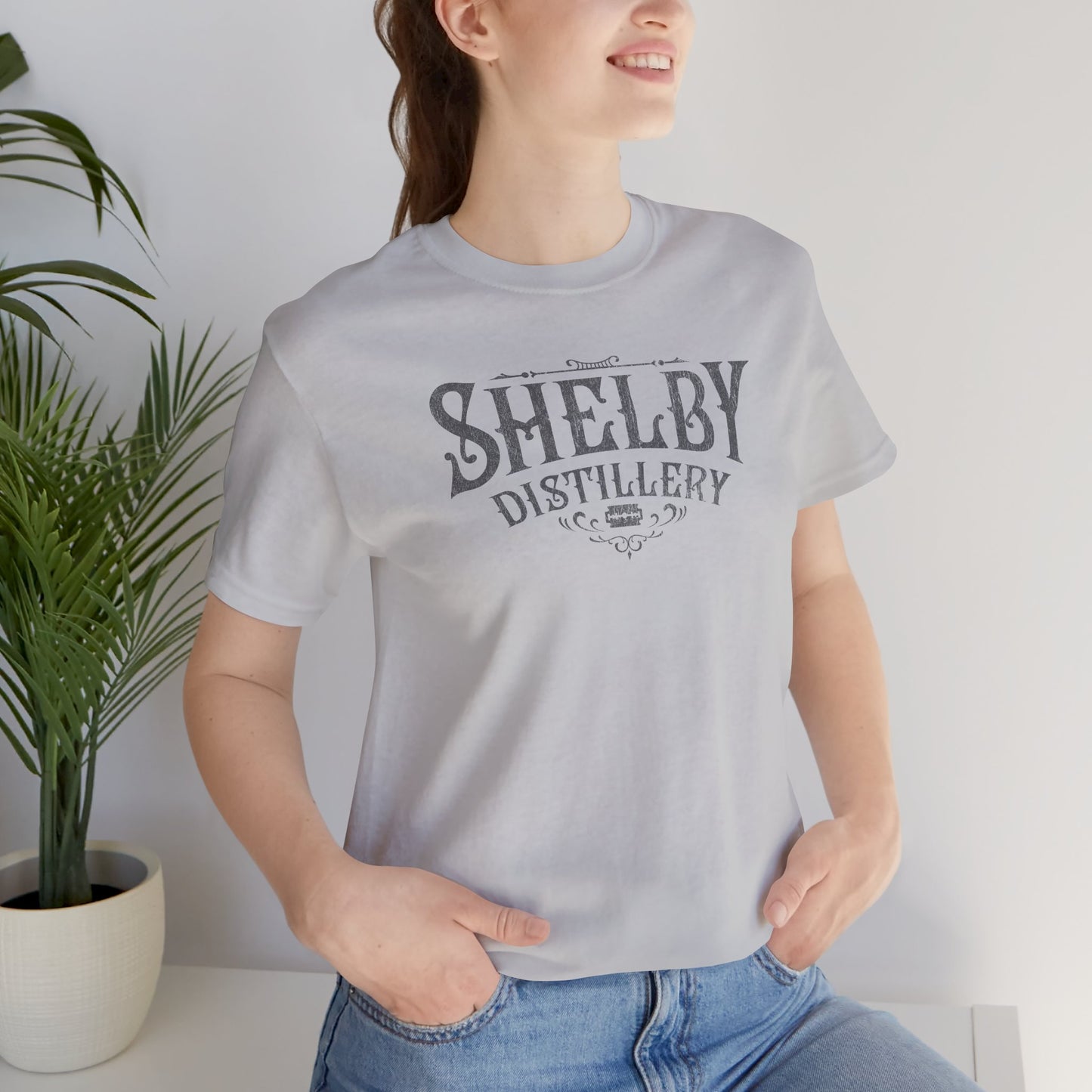 Female model wearing a solid athletic gray Soft Bella+Canvas 3001 short-sleeve t-shirt featuring the Shelby Company Limited Distillery Logo Graphic inspired by the TV show and movie Peaky Blinders in a vintage distressed style, sold by ScriptedBrand.