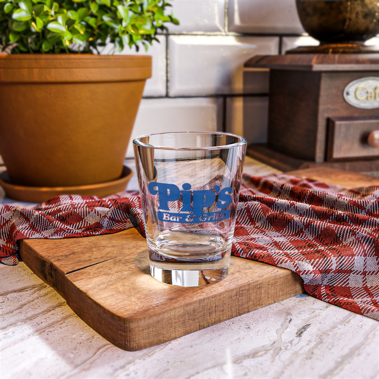 Mockup of a 1.5 oz ounce clear shot glass featuring the Pips Bar and Grill logo from the Apple TV show Severance printed on one side, shown in a kitchen setting.