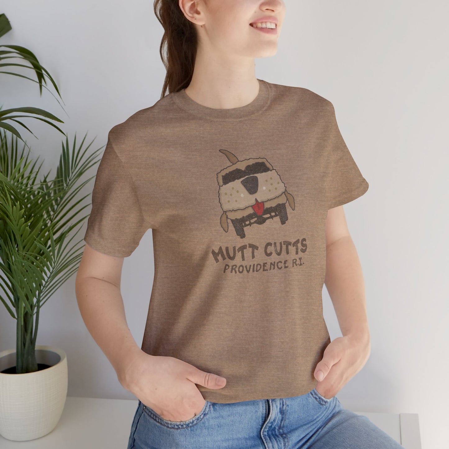 Female model wearing a heather tan colored Soft unisex Bella+Canvas 3001 short-sleeve t-shirt featuring a Mutt Cutts logo graphic inspired by the movie Dumb and Dumber, illustrated by ScriptedBrand in a vintage distressed style.