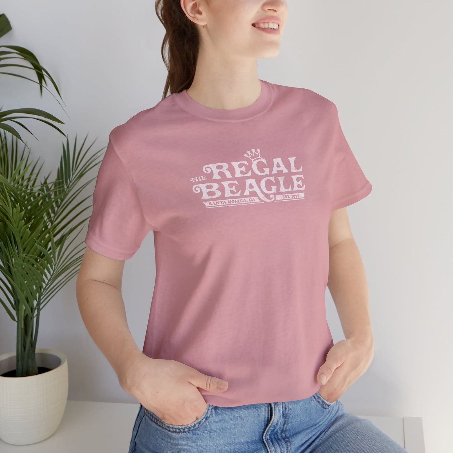 Female model wearing an orchid pink colored soft Bella+Canvas 3001 short-sleeve t-shirt featuring the Regal Beagle logo from the TV Show Threes Company in a vintage distressed style.