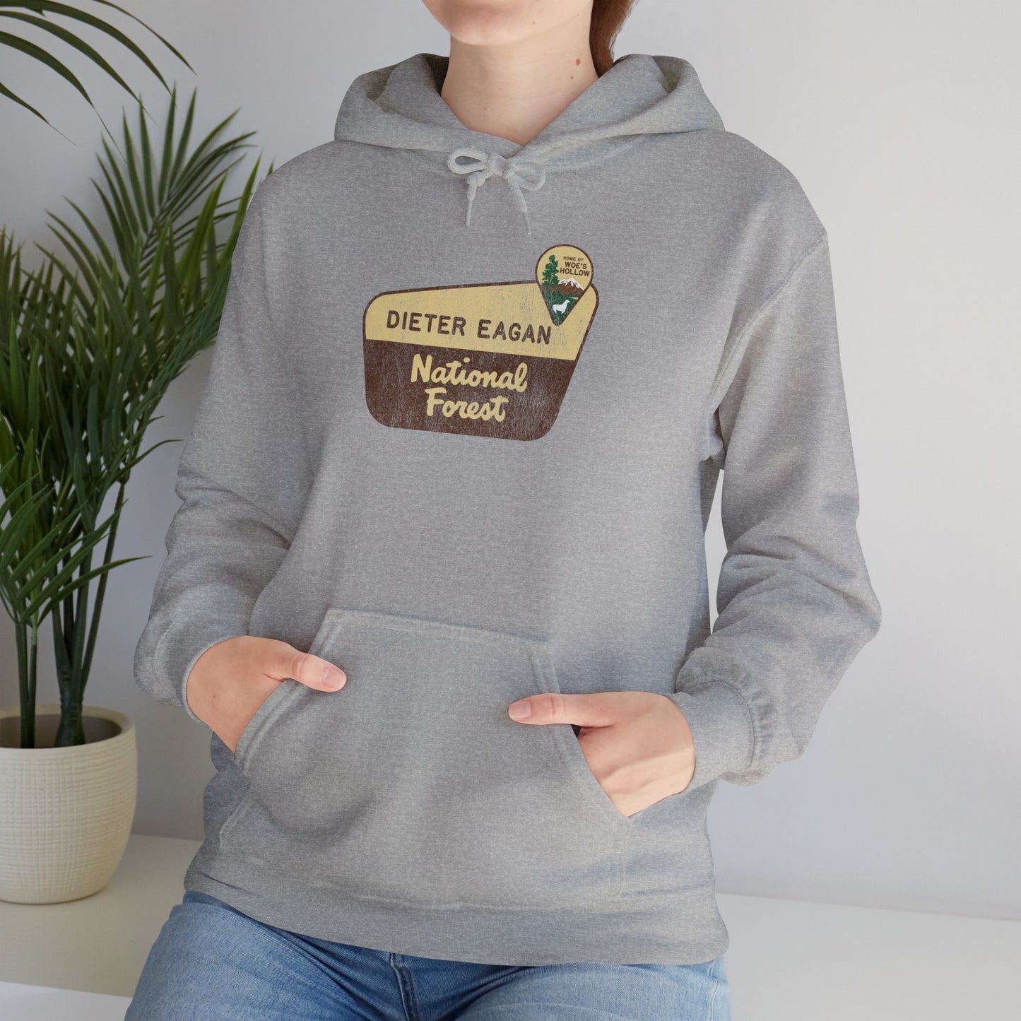 Female model wearing a Sport Heather Gray colored Soft Unisex Hoodie Heavy Blend Hooded Sweatshirt featuring the logo graphic of the Dieter Eagan National Forest inspired by the TV show Severance in a vintage distressed ink-stamped worn-in style.