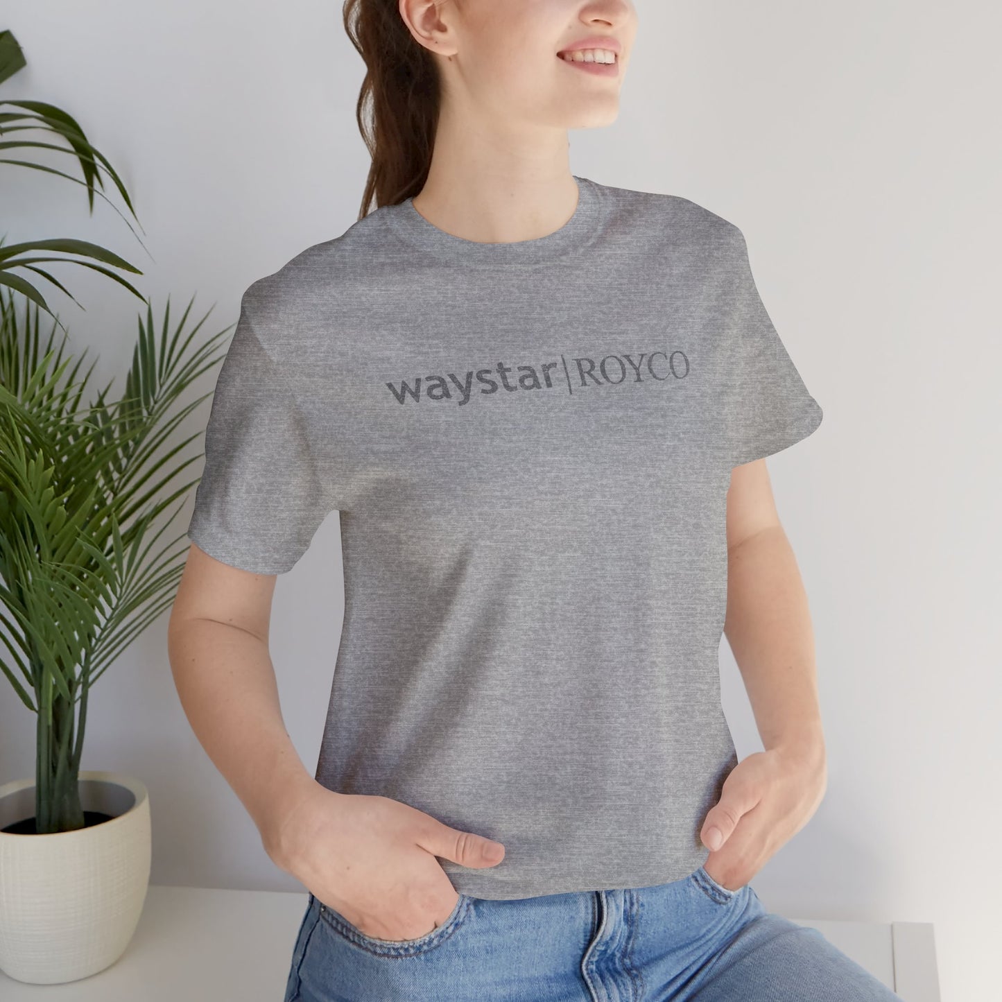 Female model wearing an athletic heather grey colored soft Bella+Canvas 3001 short-sleeve t-shirt featuring the Waystar Royco logo from the TV show Succession in a vintage distressed style, sold by ScriptedBrand.