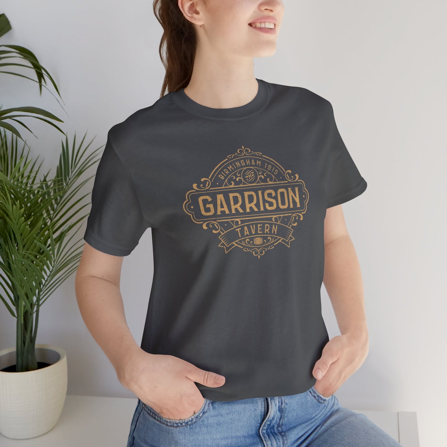 Female model wearing a asphalt gray soft Bella+Canvas 3001 short-sleeve t-shirt featuring the Garrison Tavern Bar Logo Graphic inspired by the TV show and movie Peaky Blinders in a vintage distressed style, sold by ScriptedBrand.
