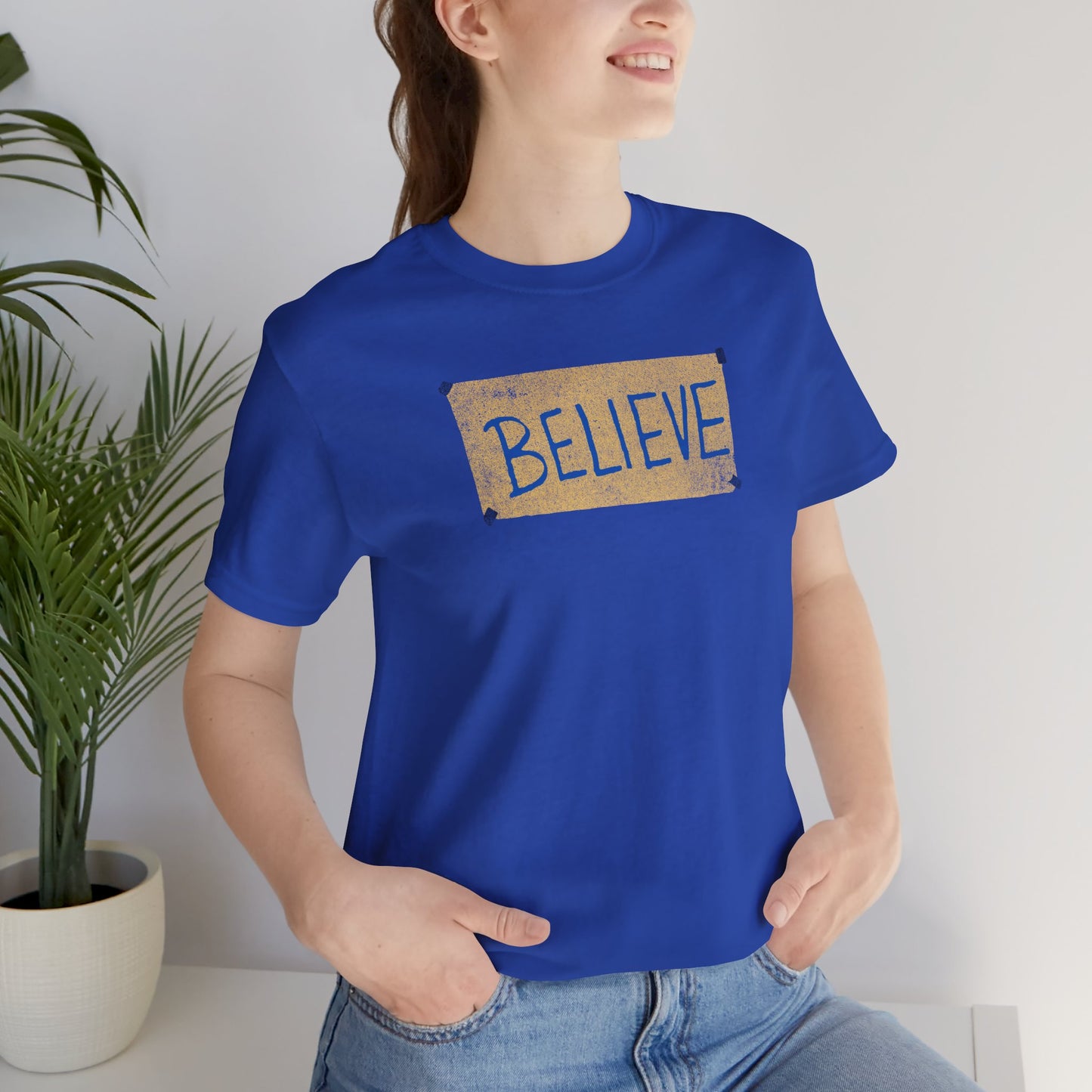 Female model wearing a royal blue colored soft Bella+Canvas 3001 short-sleeve t-shirt featuring a logo graphic of the BELIEVE sign Ted Lasso hung in the fictional English soccer team AFC Richmond locker room, inspired by the TV show Ted Lasso in a vintage distressed style, sold by ScriptedBrand.