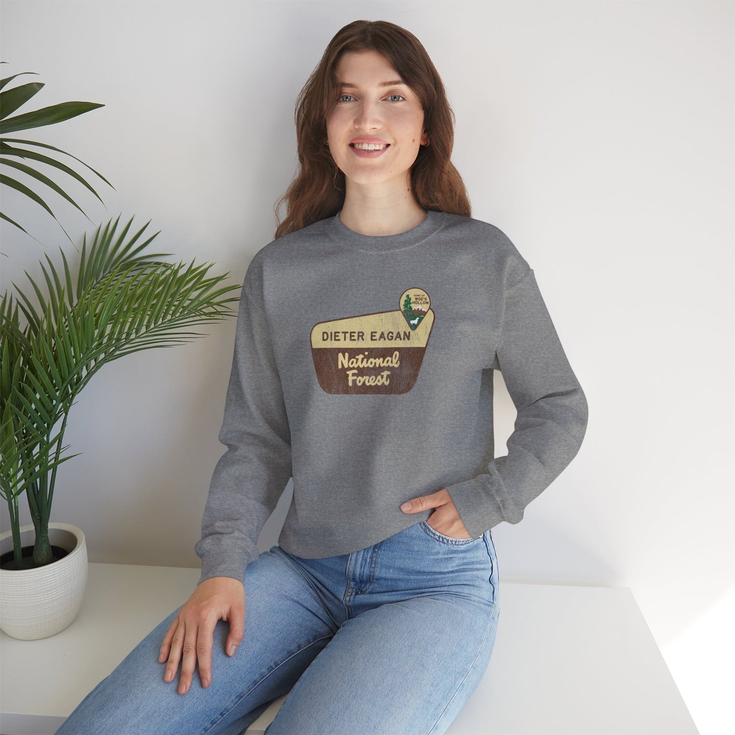 Female model wearing a dark gray heather Soft Unisex Gildan 18000 sweatshirt featuring a logo graphic of the Dieter Eagan National Forest sign designed by ScriptedBrand, inspired by the TV show Severance in a vintage distressed style.