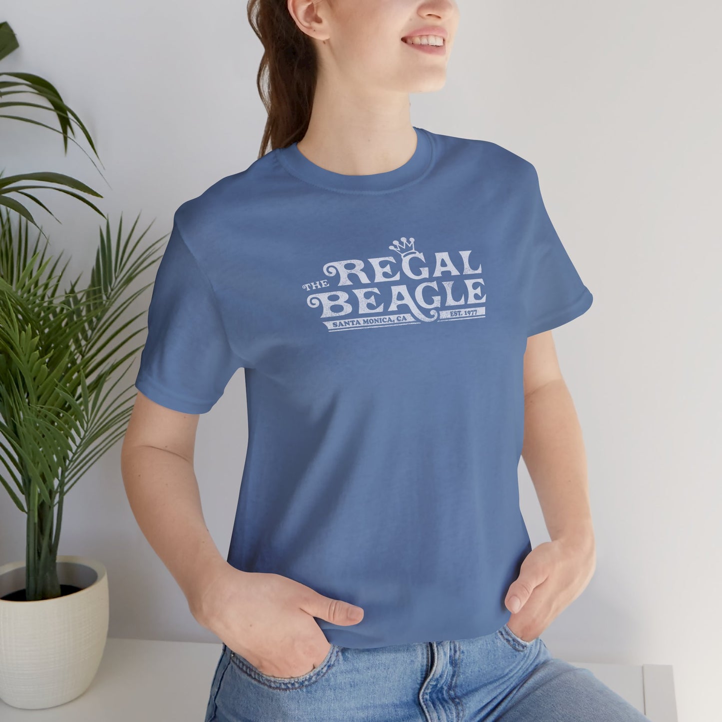 Female model wearing a Lavender Blue colored soft Bella+Canvas 3001 short-sleeve t-shirt featuring the Regal Beagle logo from the TV Show Threes Company in a vintage distressed style.