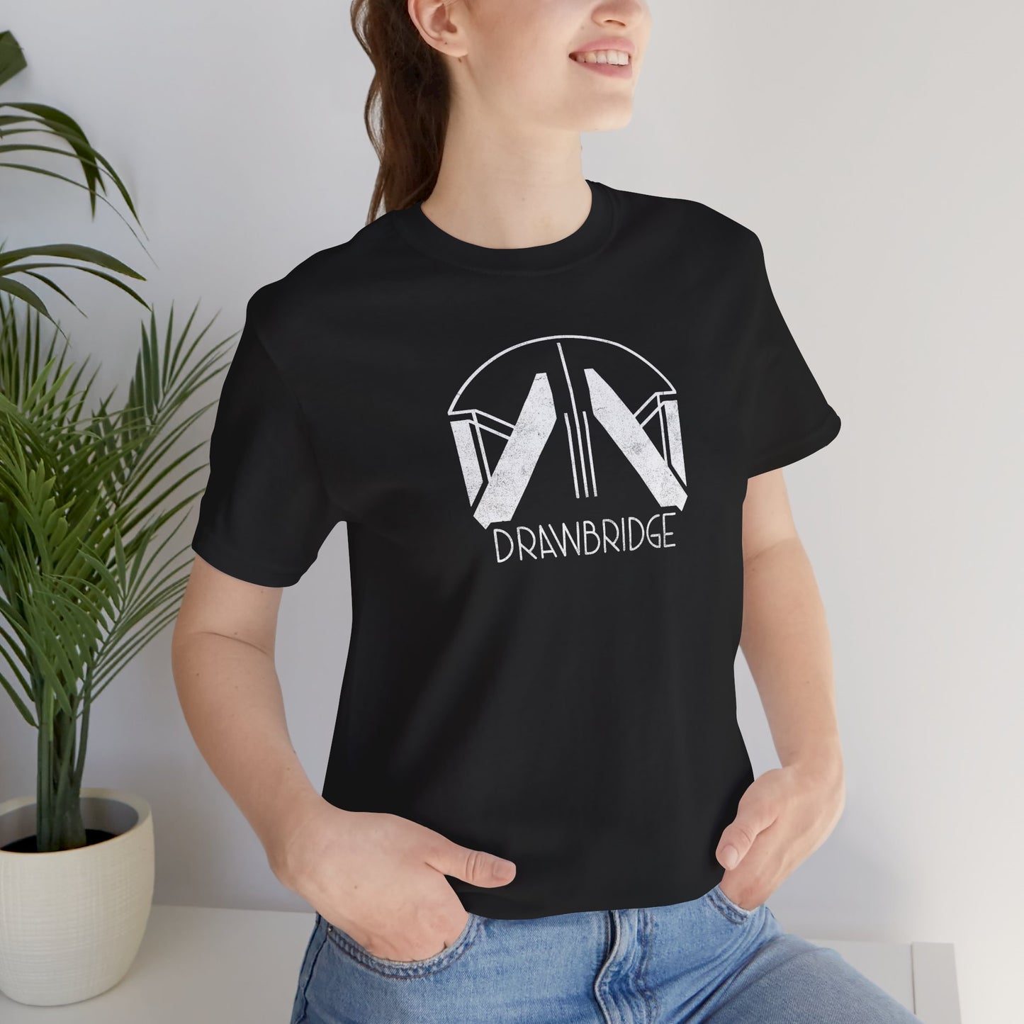 Female model wearing a black colored soft Bella+Canvas 3001 short-sleeve t-shirt featuring the Drawbridge logo from the video game Death Stranding 2, in a vintage distressed style, sold by ScriptedBrand.