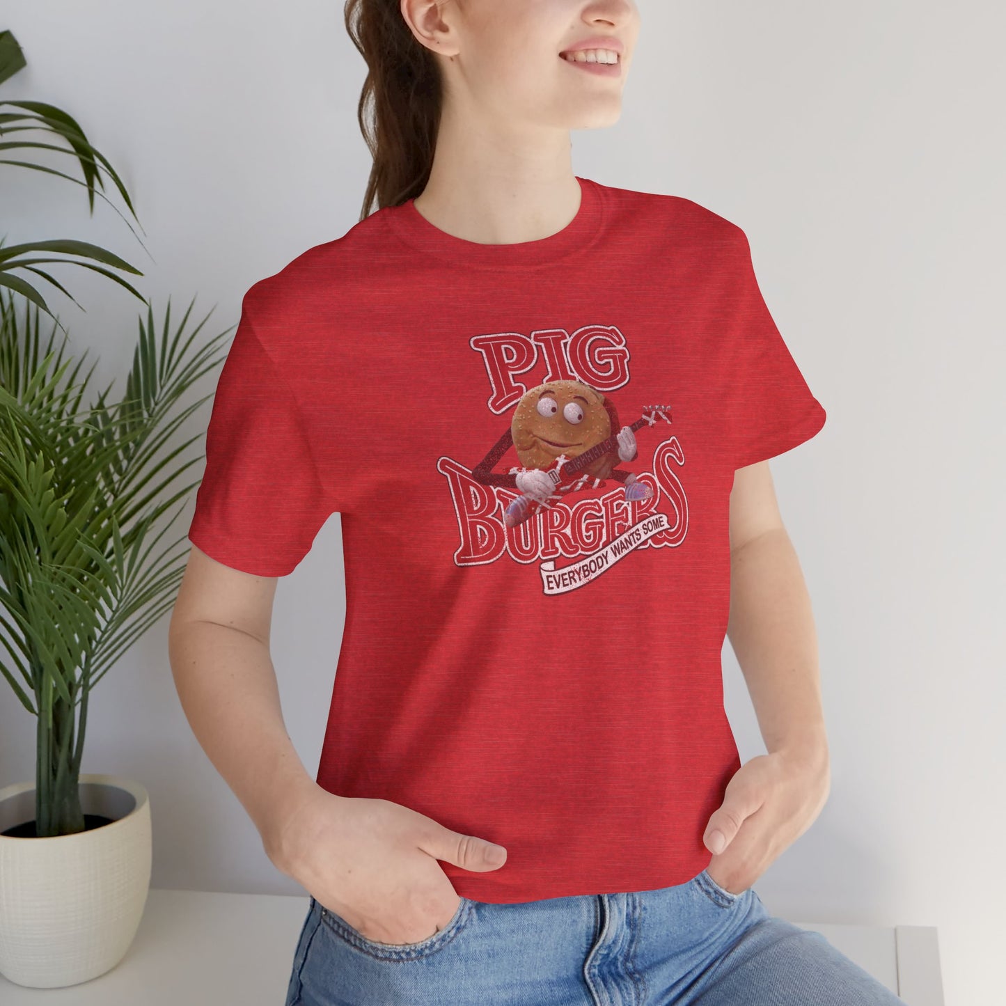 Female model wearing a heather red colored soft Bella+Canvas 3001 short-sleeve t-shirt featuring a graphic of the fictional Pig Burgers logo from the movie Better Off Dead, along with the Van Halen singing hamburger playing guitar, with the small subhead phrase EVERYBODY WANTS SOME. The graphic is designed in a vintage distressed style, sold by ScriptedBrand.