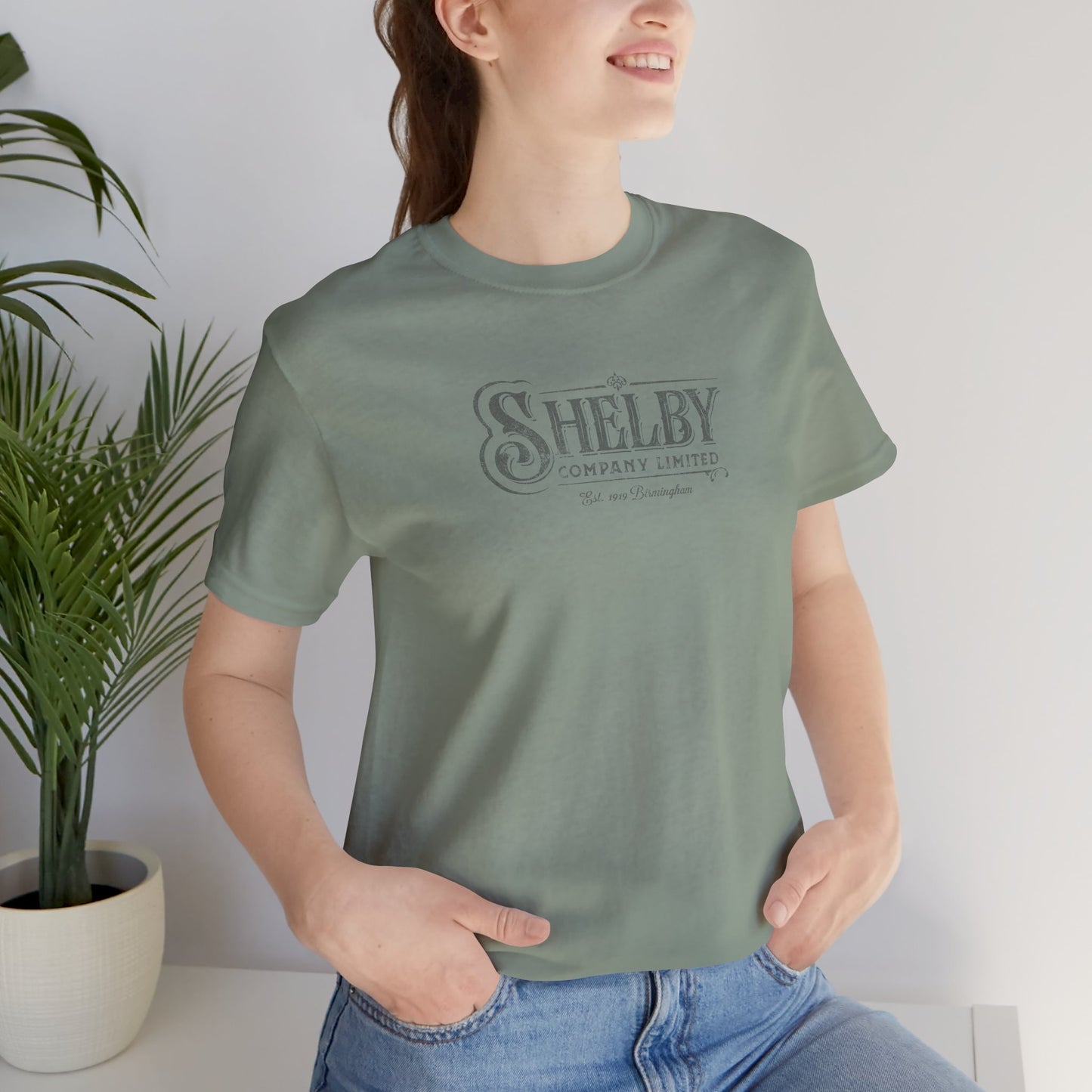 Female model wearing sage green soft short-sleeve Bella+Canvas 3001 t-shirt featuring the Shelby Company Limited logo from the show Peaky Blinders in a vintage distressed style, sold by ScriptedBrand.