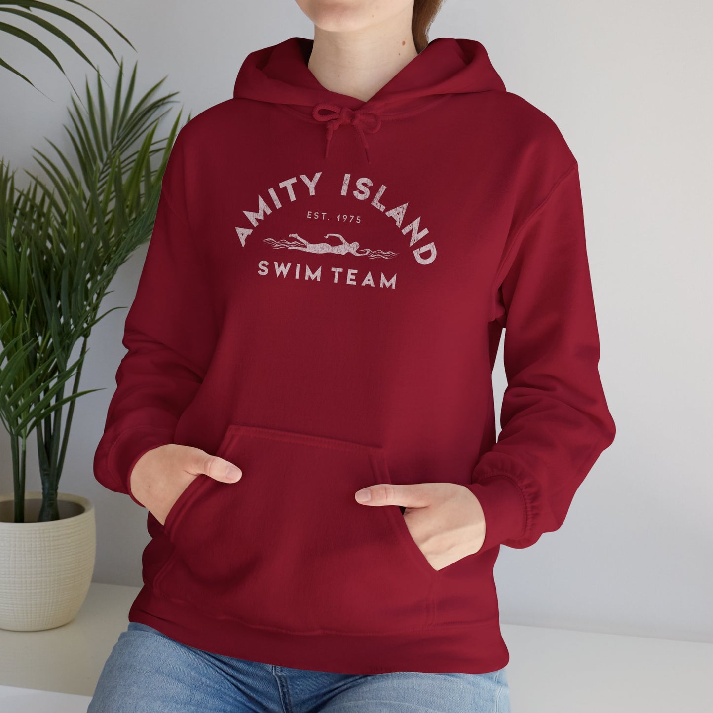 Female model wearing a lifeguard cardinal red colored Soft Unisex Hoodie Heavy Blend Hooded Sweatshirt featuring an Amity Island Swim Team logo graphic, designed by ScriptedBrand, and inspired by the movie Jaws in a vintage distressed style.