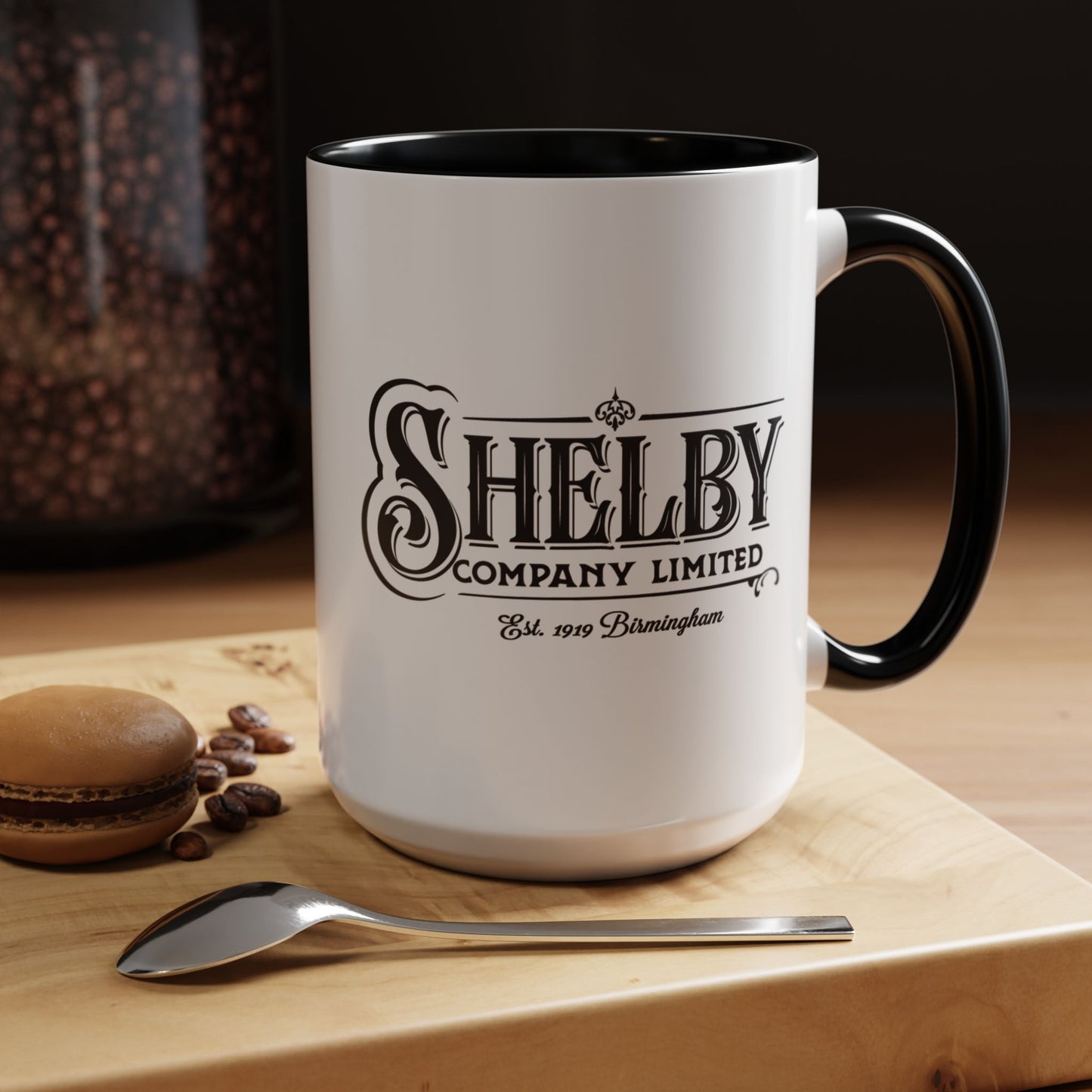 Front view of a 15 oz White Ceramic Mug with black handle/interior featuring the fictional Shelby Company Limited Logo from the TV show Peaky Blinders printed on both sides. Graphic is designed by ScriptedBrand and includes the text "Est. 1919 Birmingham". Mug is shown placed in a kitchen setting.