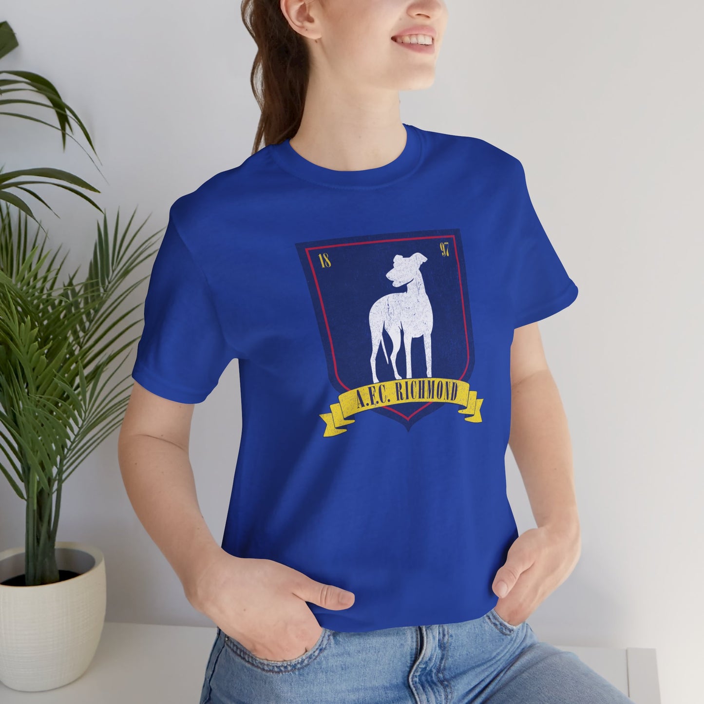 Female model wearing a royal blue soft Bella+Canvas 3001 short-sleeve t-shirt featuring the logo graphic of the fictional English soccer team AFC Richmond inspired by the TV show Ted Lasso in a vintage distressed style, sold by ScriptedBrand.