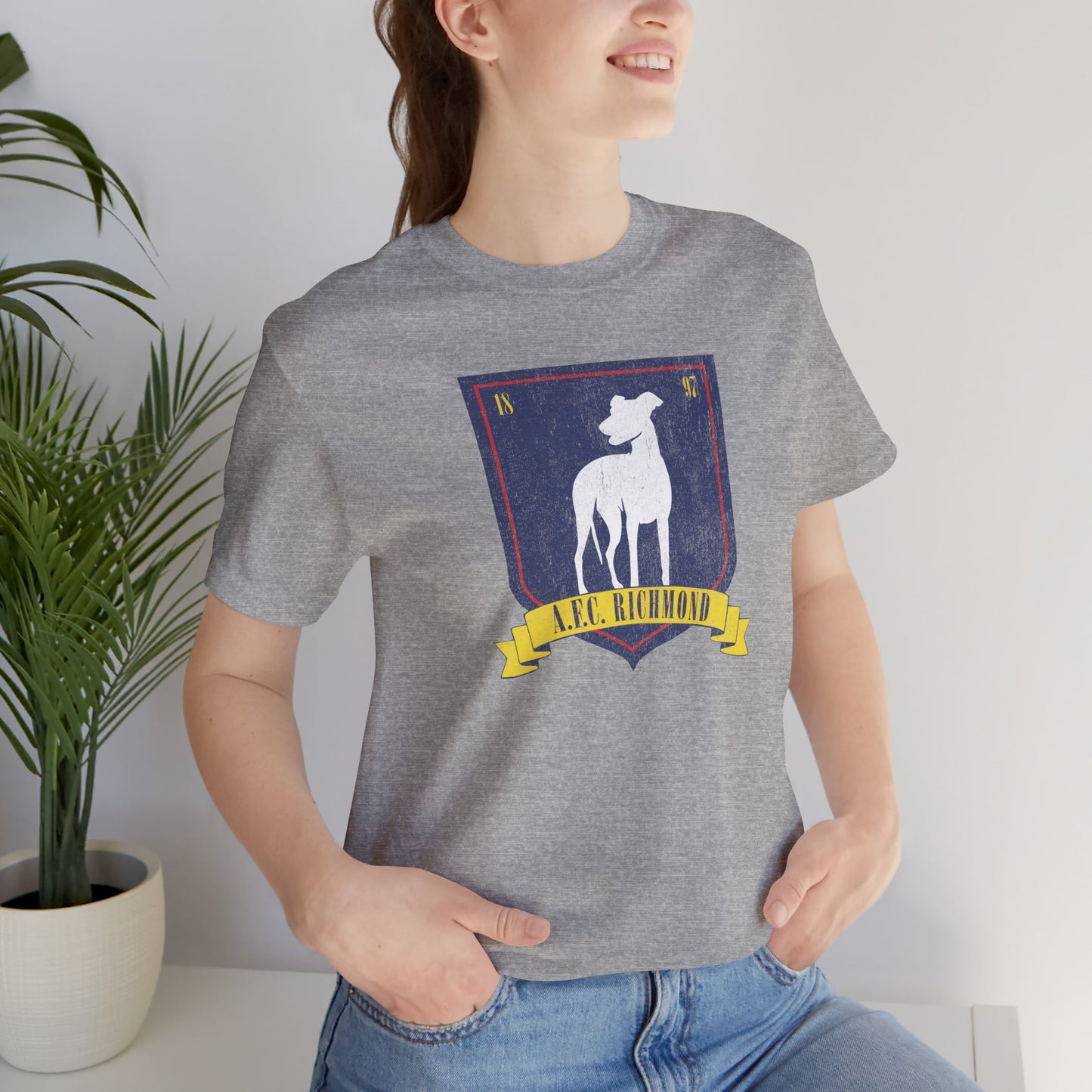 Female model wearing an athletic heather grey soft Bella+Canvas 3001 short-sleeve t-shirt featuring the logo graphic of the fictional English soccer team AFC Richmond inspired by the TV show Ted Lasso in a vintage distressed style, sold by ScriptedBrand.