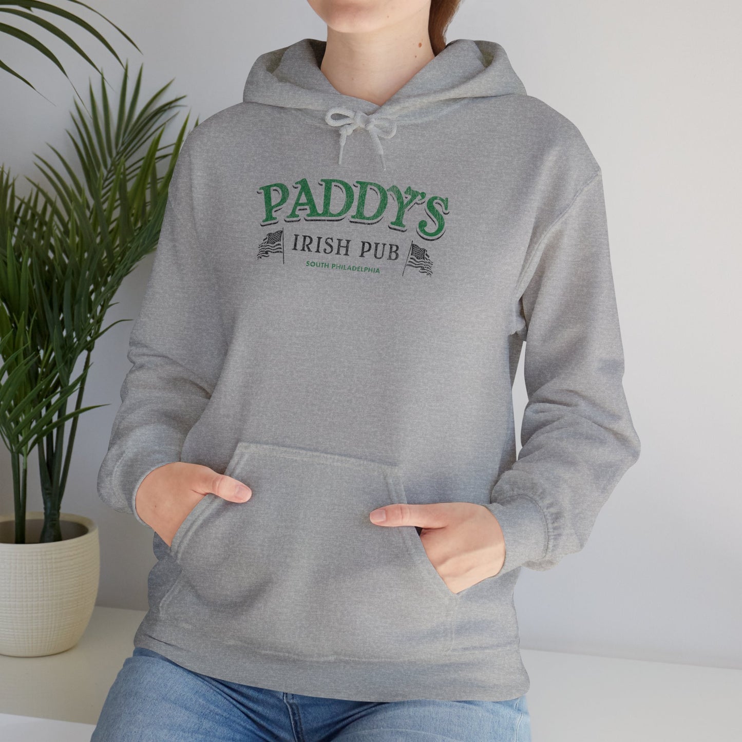 Female model wearing a sport heather grey colored soft unisex Hoodie Heavy Blend Hooded Sweatshirt featuring the green Paddy's Pub logo graphic from the TV Show It's Always Sunny in Philadelphia, in a vintage distressed style, sold by ScriptedBrand.