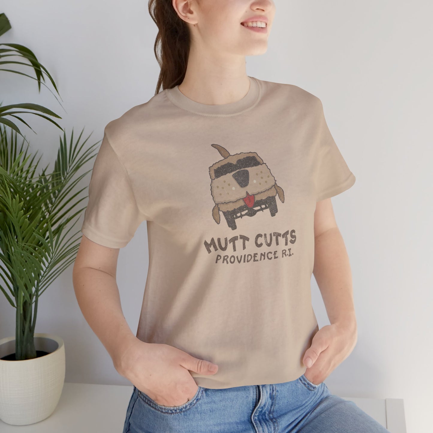 Female model wearing a tan colored Soft unisex Bella+Canvas 3001 short-sleeve t-shirt featuring a Mutt Cutts logo graphic inspired by the movie Dumb and Dumber, illustrated by ScriptedBrand in a vintage distressed style.
