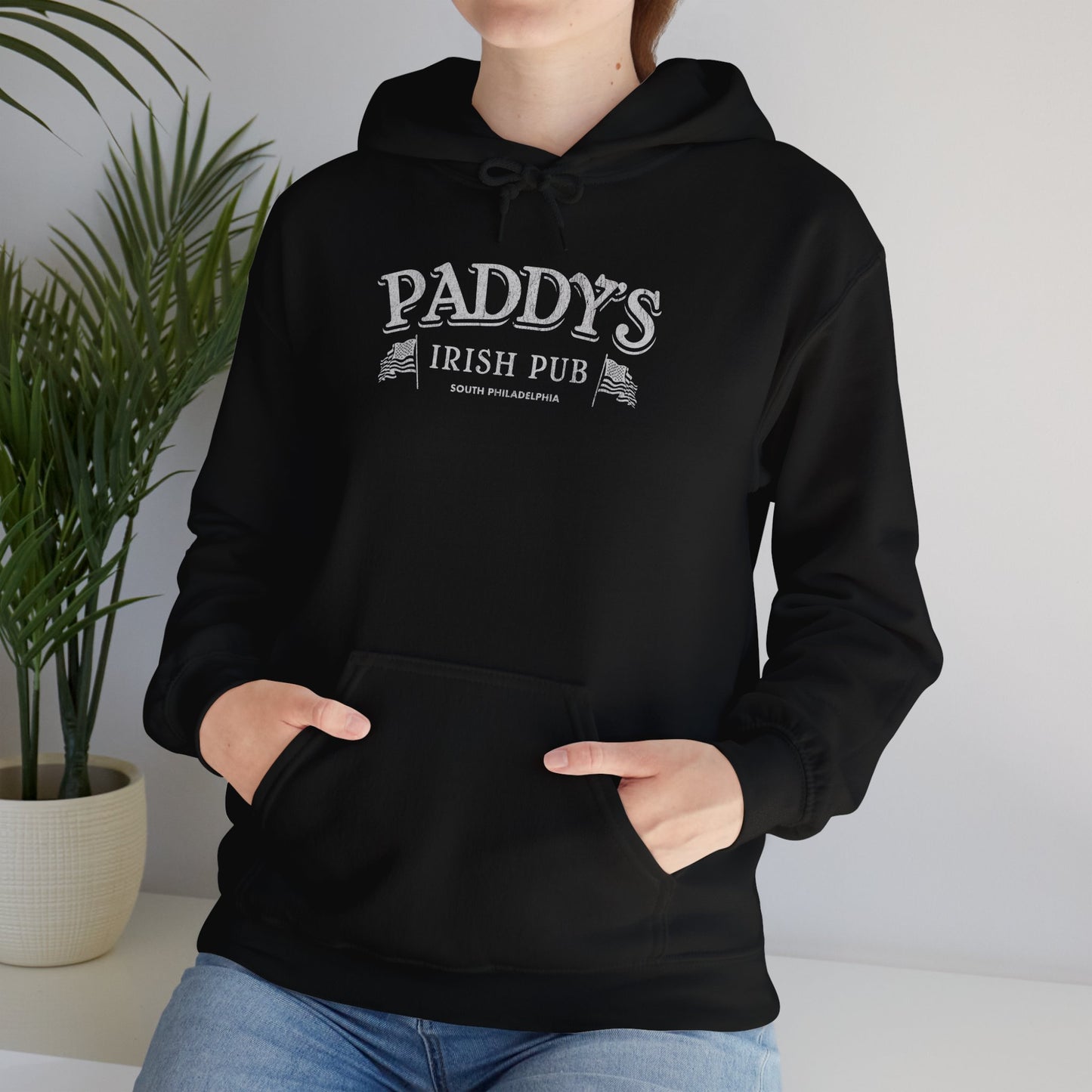 Female model wearing a black colored soft unisex Hoodie Heavy Blend Hooded Sweatshirt featuring the Paddy's Pub logo graphic from the TV Show It's Always Sunny in Philadelphia, in a vintage distressed style, sold by ScriptedBrand.