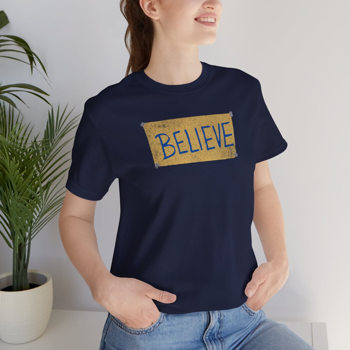 Female model wearing a navy blue colored soft Bella+Canvas 3001 short-sleeve t-shirt featuring a logo graphic of the BELIEVE sign Ted Lasso hung in the fictional English soccer team AFC Richmond locker room, inspired by the TV show Ted Lasso in a vintage distressed style, sold by ScriptedBrand.