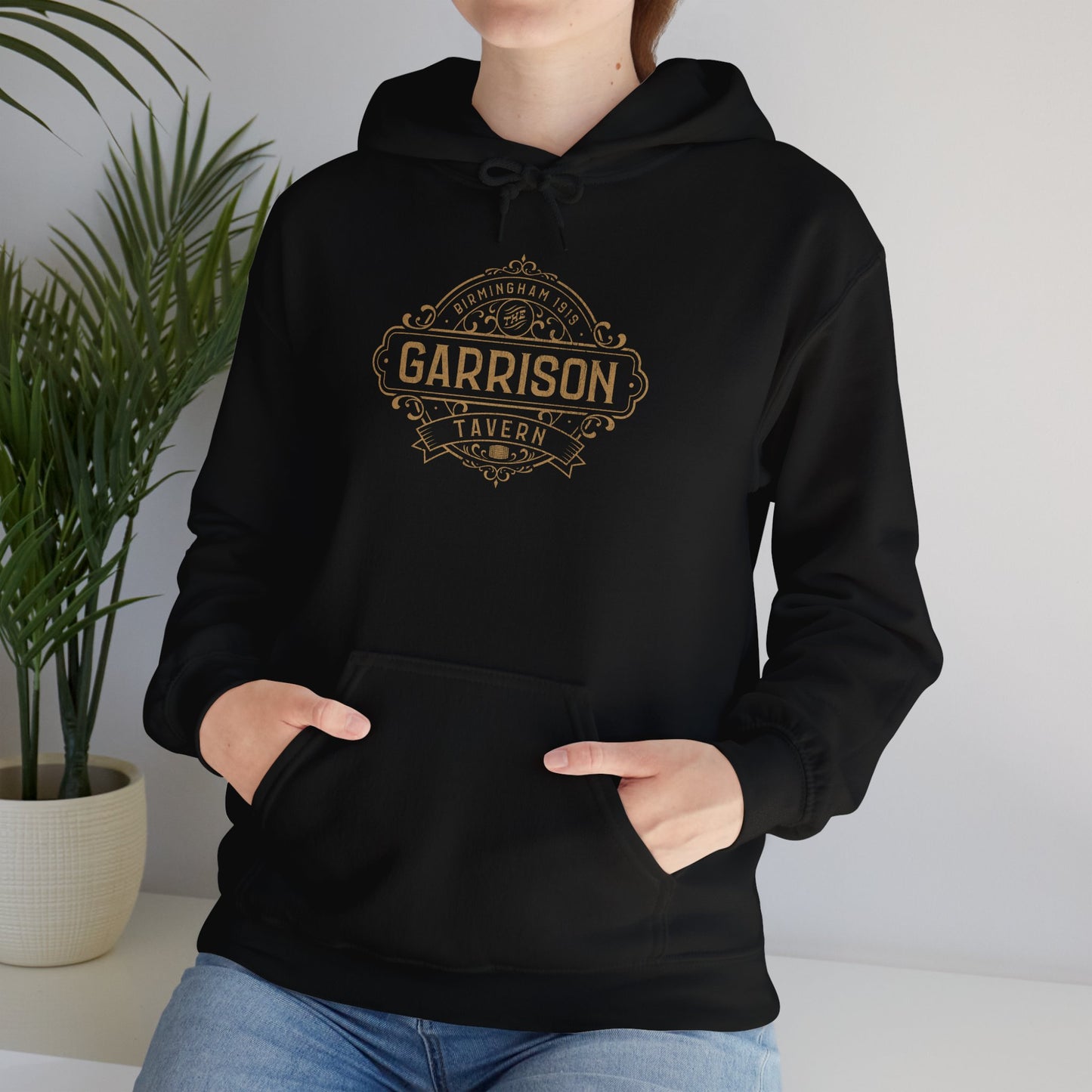 Female model wearing a black Soft Unisex Hoodie Heavy Blend Hooded Sweatshirts featuring the Garrison Tavern Bar Logo Graphic inspired by the TV show and movie Peaky Blinders in a vintage distressed style sold by ScriptedBrand.