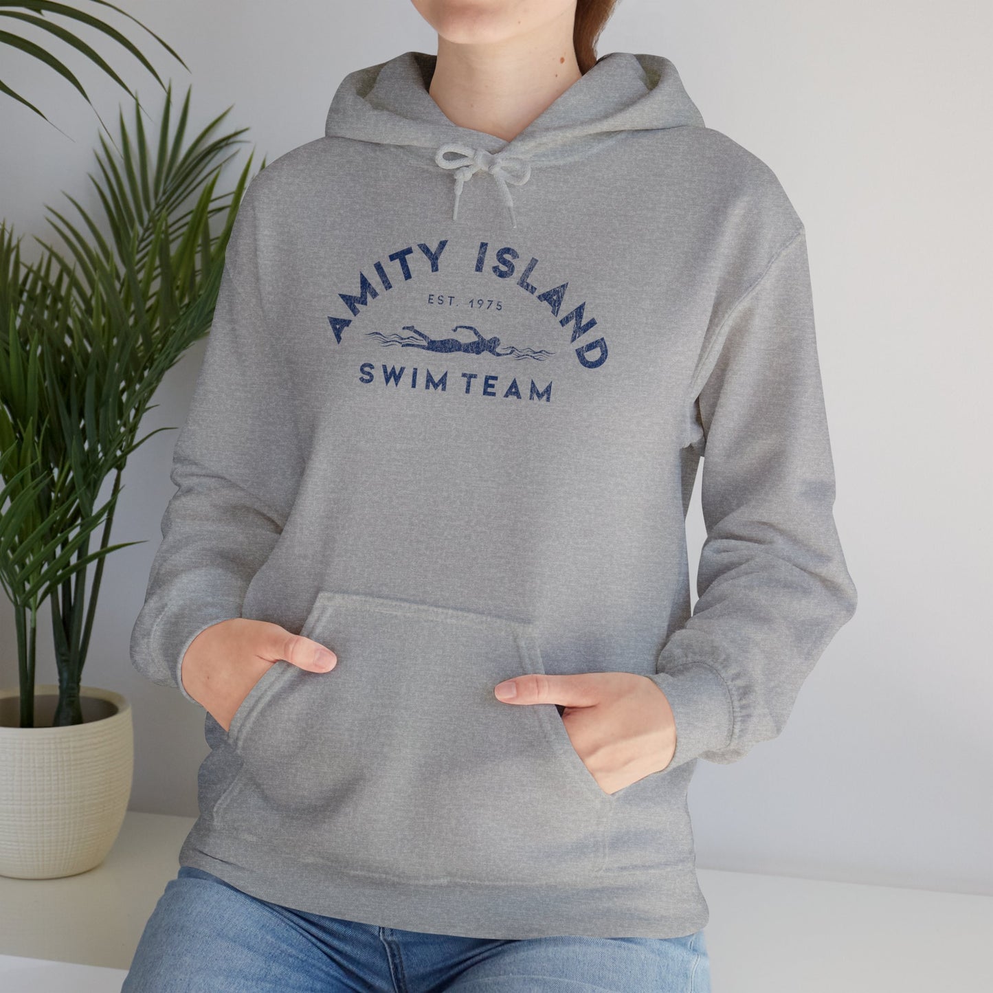 Female model wearing a sport heather grey colored Soft Unisex Hoodie Heavy Blend Hooded Sweatshirt featuring an Amity Island Swim Team logo graphic, designed by ScriptedBrand, and inspired by the movie Jaws in a vintage distressed style.