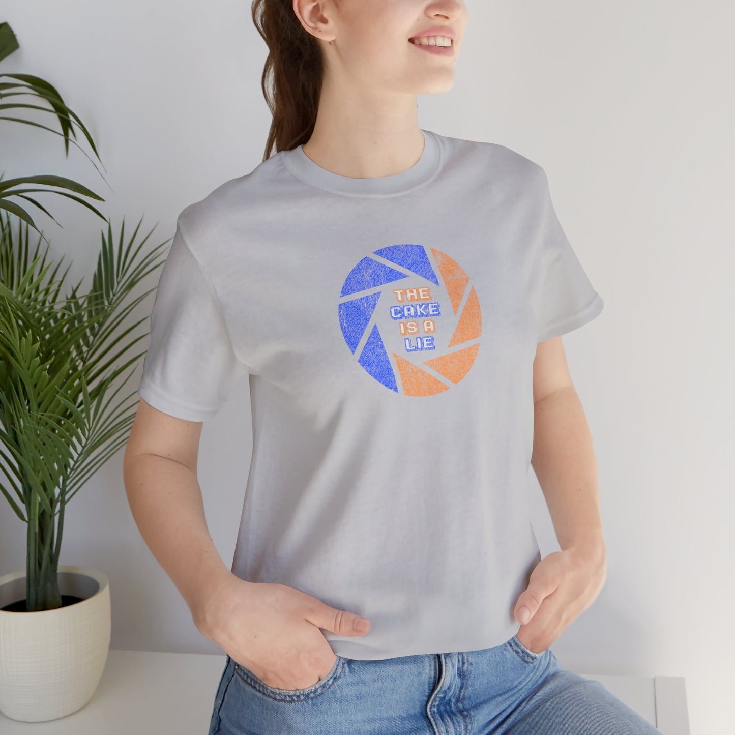 Female model wearing a solid athletic grey colored soft Bella+Canvas 3001 short-sleeve t-shirt featuring the phrase The Cake Is a Lie, along with the Aperture Laboratories shutter logo from the video game Portal and Portal 2 in a vintage distressed style, sold by ScriptedBrand.