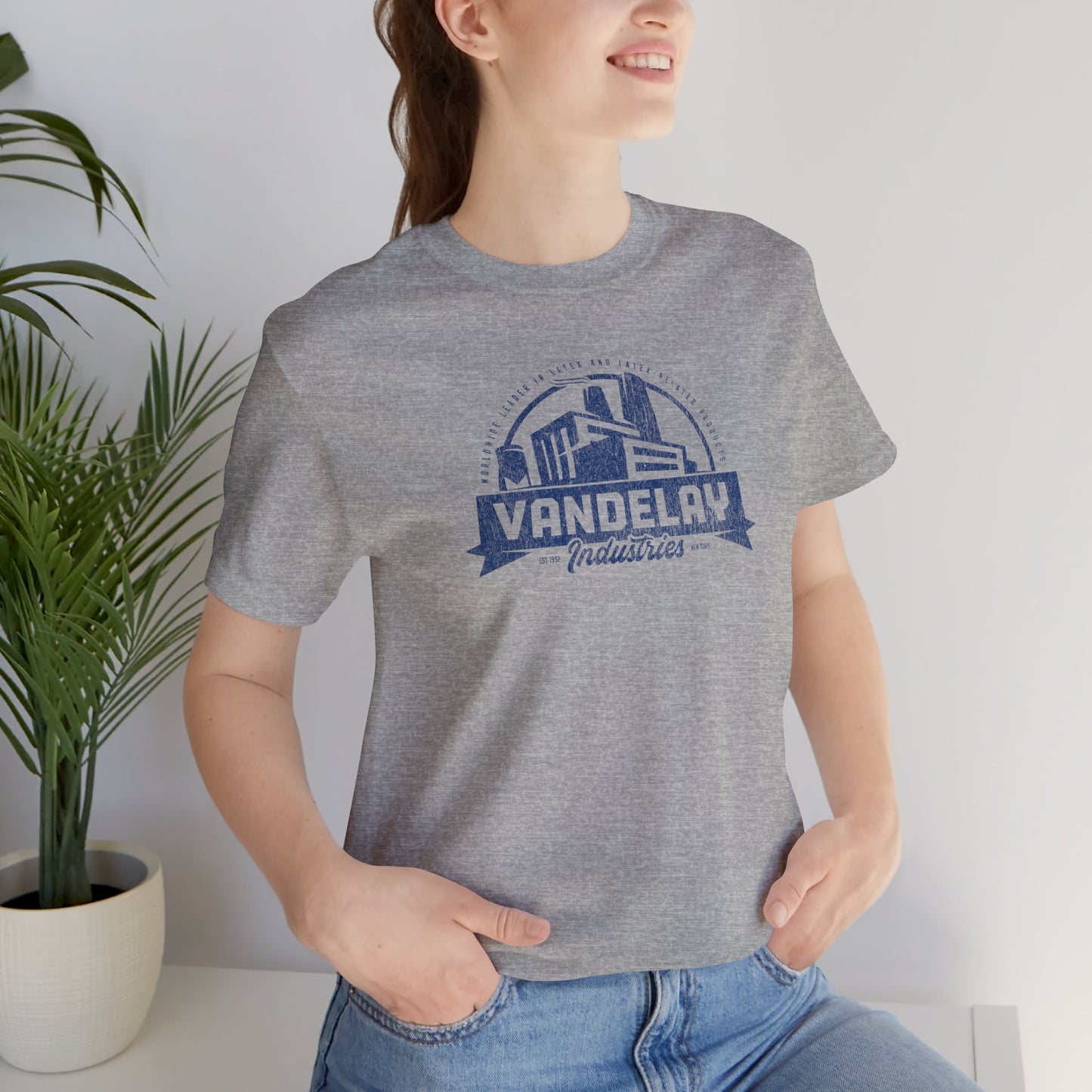 Female model wearing an Athletic Heather Grey Soft Bella+Canvas 3001 short-sleeve t-shirt tee featuring a Vandelay Industries logo graphic, uniquely designed by ScriptedBrand in a vintage distressed style. The fictional company is part of the George Costanza storyline in the TV show Seinfeld.