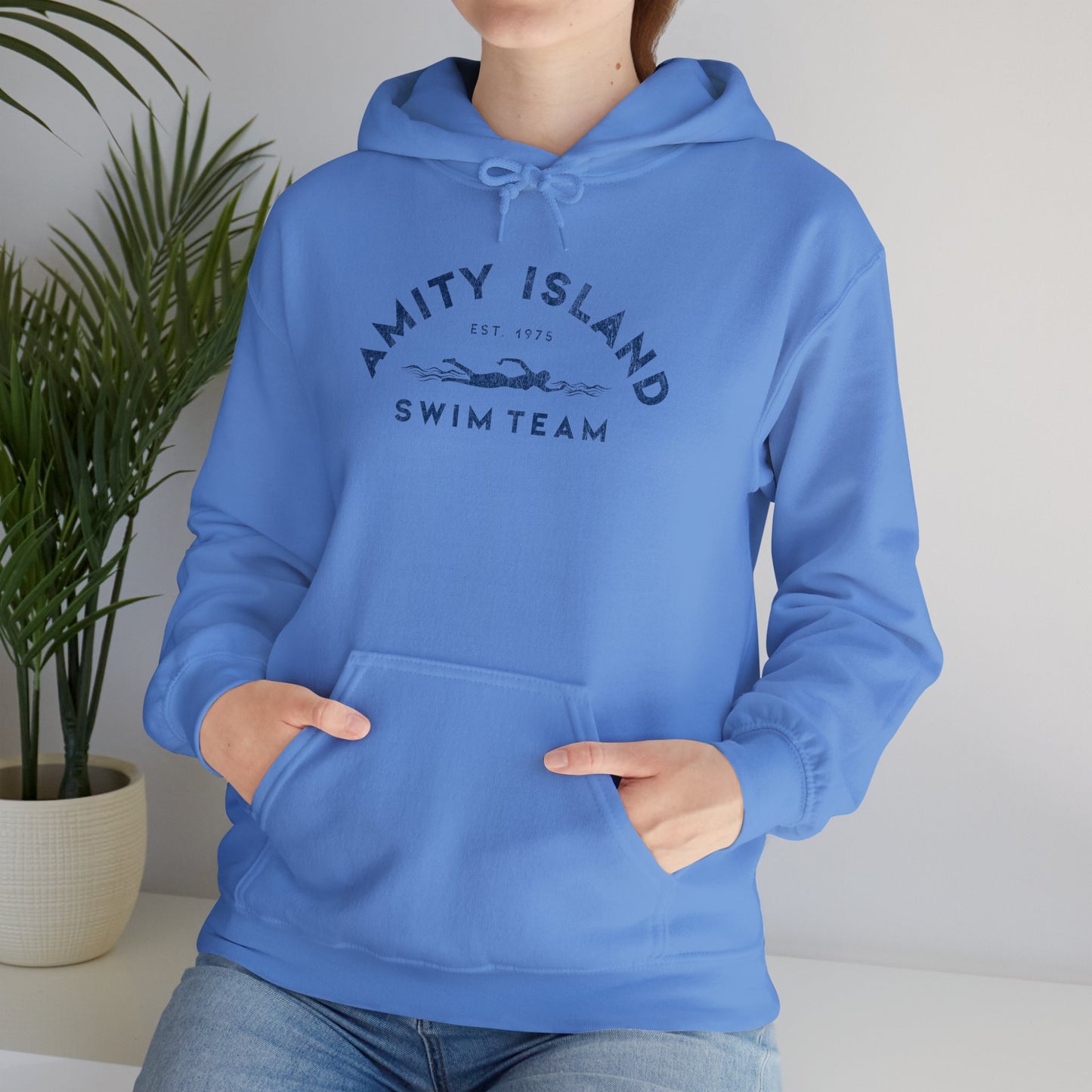 Female model wearing a Carolina blue colored Soft Unisex Hoodie Heavy Blend Hooded Sweatshirt featuring an Amity Island Swim Team logo graphic, designed by ScriptedBrand, and inspired by the movie Jaws in a vintage distressed style.