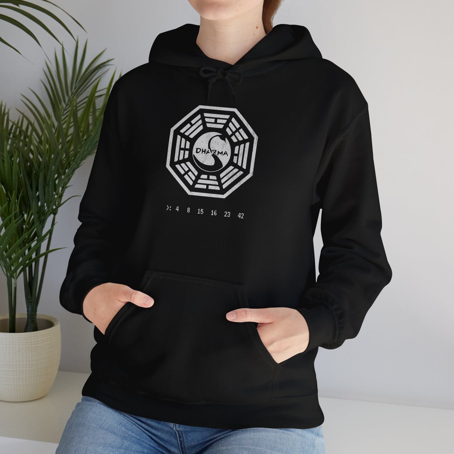 Female model wearing a Black Soft Unisex Hoodie Heavy Blend Hooded Sweatshirt featuring the Dharma Initiative swan logo graphic from the TV show LOST in a vintage distressed ink-stamped worn-in style.