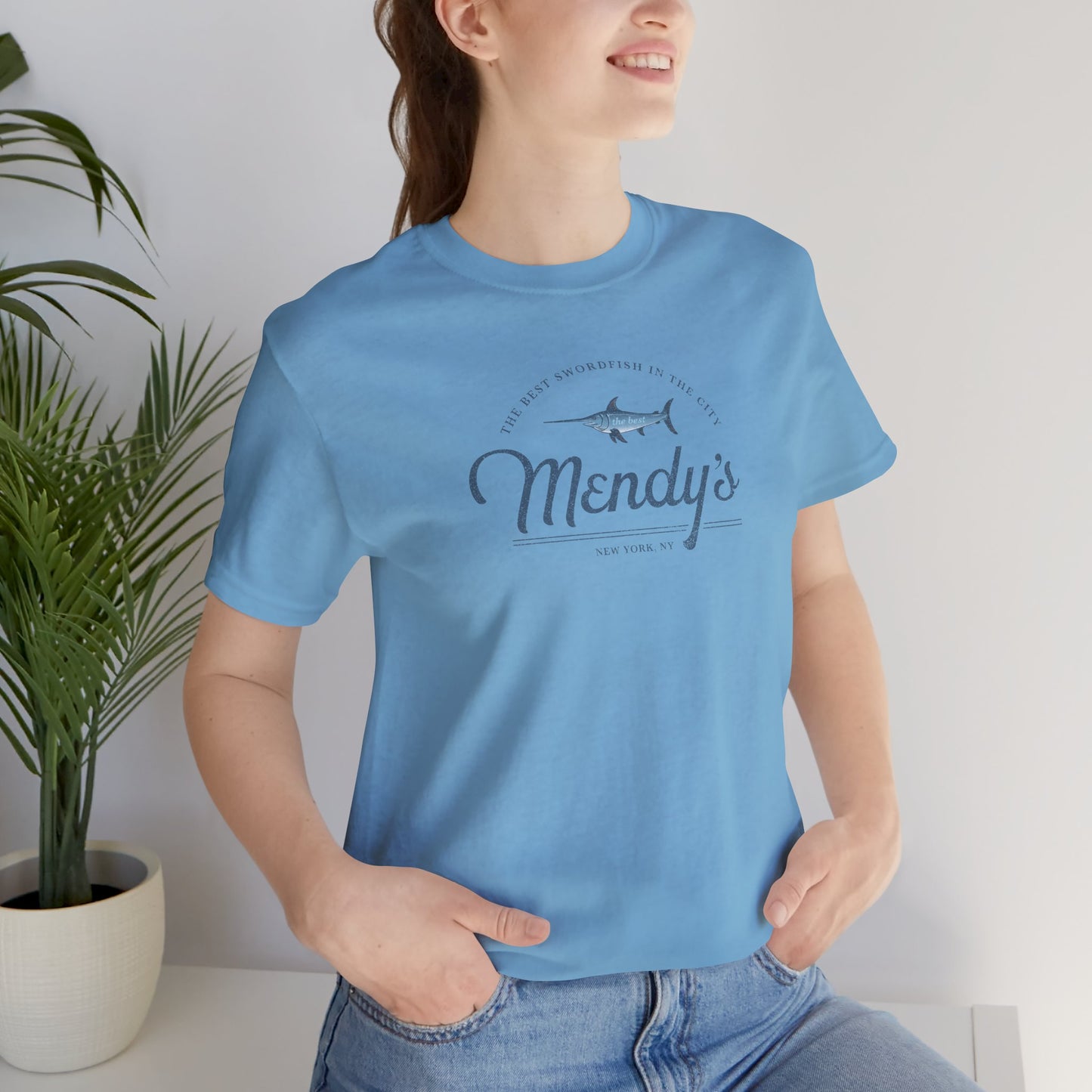 Female model wearing an ocean blue colored soft unisex Bella+Canvas 3001 short-sleeve t-shirt featuring a unique Mendy's Restaurant logo graphic, designed by ScriptedBrand in a vintage distressed style, and inspired by the TV show Seinfeld.