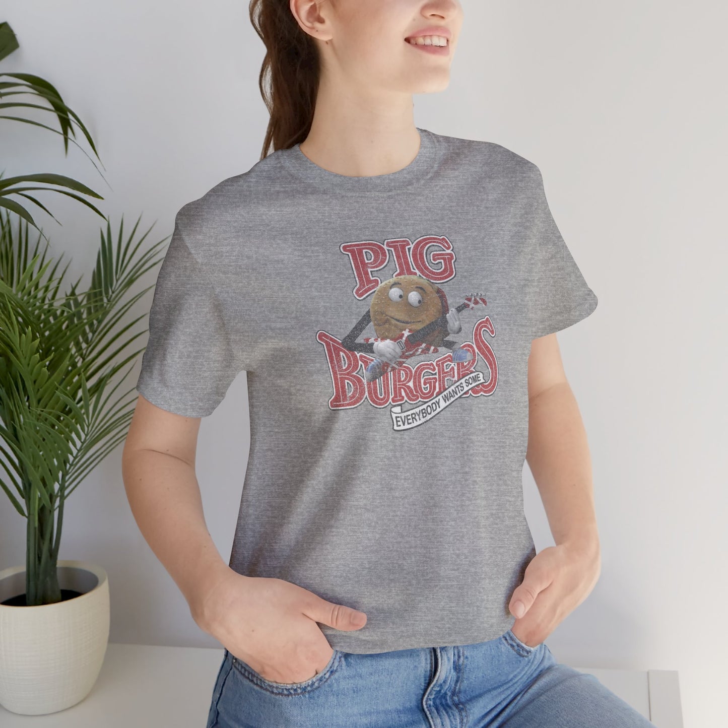 Female model wearing an athletic heather grey colored soft Bella+Canvas 3001 short-sleeve t-shirt featuring a graphic of the fictional Pig Burgers logo from the movie Better Off Dead, along with the Van Halen singing hamburger playing guitar, with the small subhead phrase EVERYBODY WANTS SOME. The graphic is designed in a vintage distressed style, sold by ScriptedBrand.