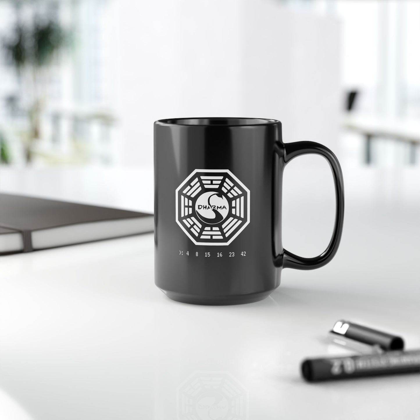 Black Ceramic Mug on a desk featuring the Dharma Initiative swan station logo from the TV show LOST, including the Hatch numbers 4 8 15 16 23 42. Graphic is printed white on both sides.