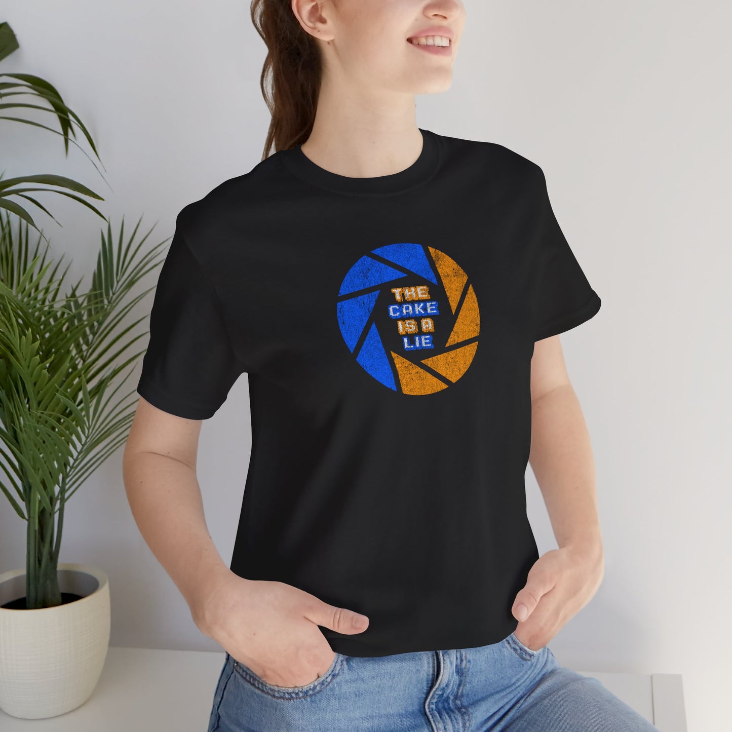 Female model wearing a black colored soft Bella+Canvas 3001 short-sleeve t-shirt featuring the phrase The Cake Is a Lie, along with the Aperture Laboratories shutter logo from the video game Portal and Portal 2 in a vintage distressed style, sold by ScriptedBrand.