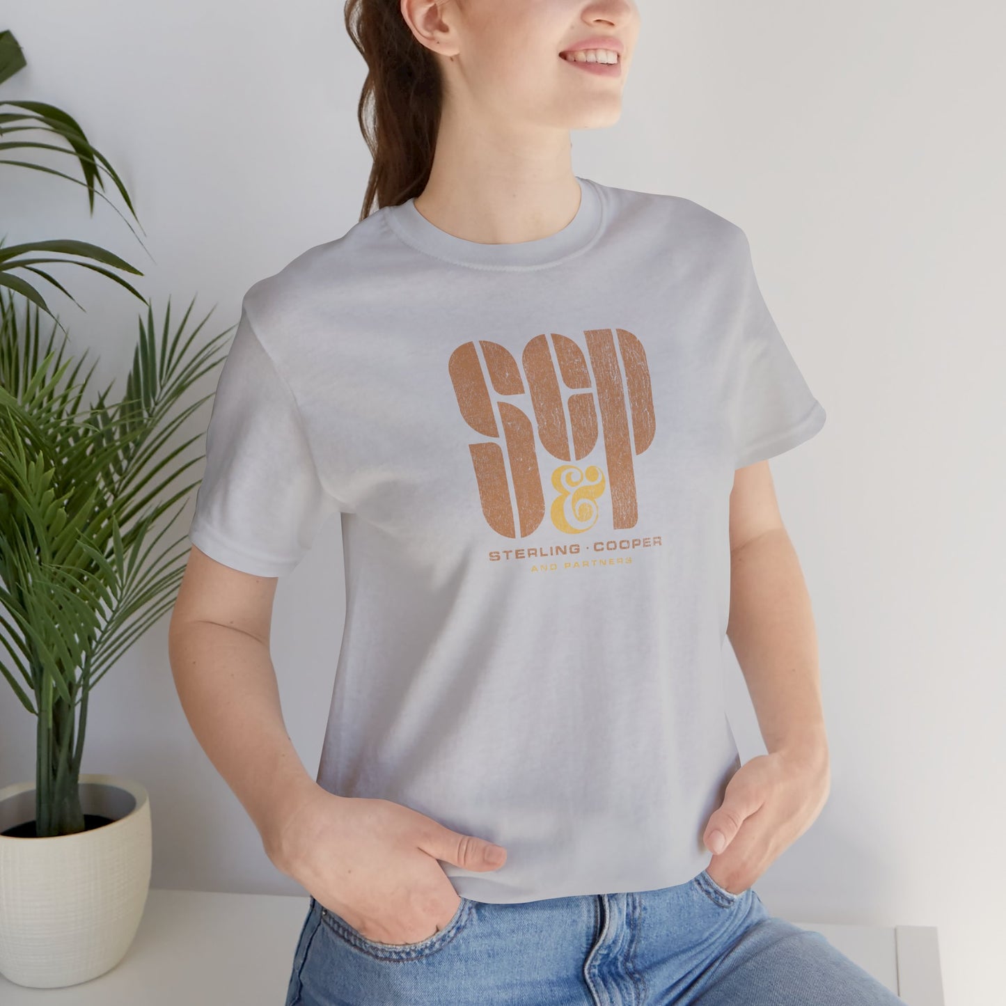 Female model wearing a solid athletic grey colored soft unisex Bella+Canvas 3001 short-sleeve t-shirt featuring the Sterling Cooper & Partners logo graphic from the TV Show Mad Men in a vintage distressed style, sold by ScriptedBrand.