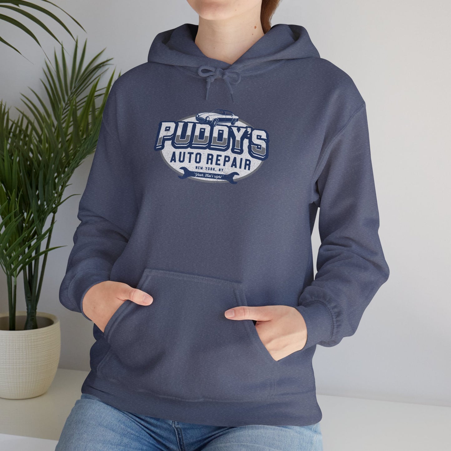 Female model wearing a heather navy blue soft Unisex Hoodie Heavy Blend Hooded Sweatshirt featuring a Puddy's Auto Repair graphic designed by ScriptedBrand inspired by the TV show Seinfeld in a vintage distressed style.