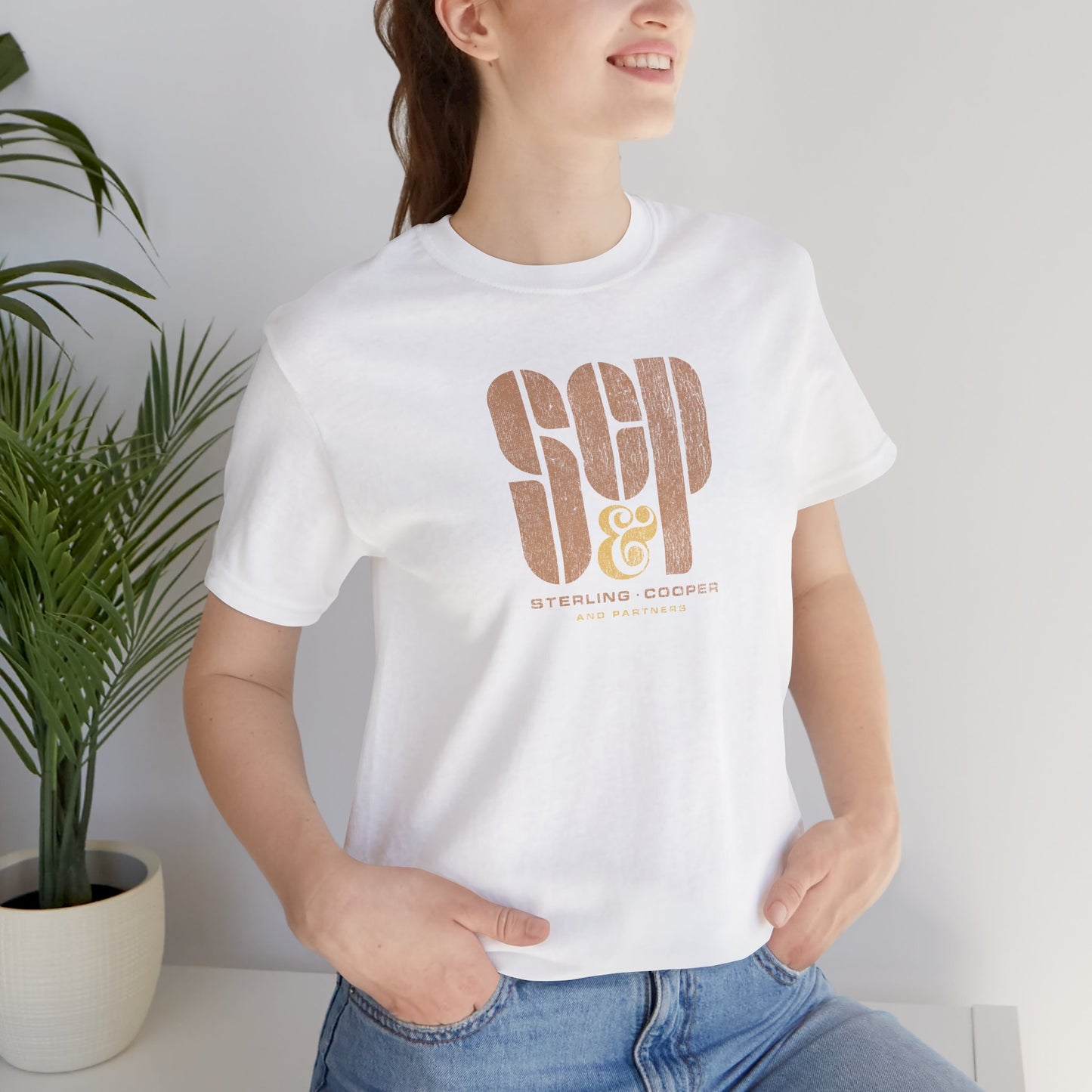 Female model wearing a white colored soft unisex Bella+Canvas 3001 short-sleeve t-shirt featuring the Sterling Cooper & Partners logo graphic from the TV Show Mad Men in a vintage distressed style, sold by ScriptedBrand.