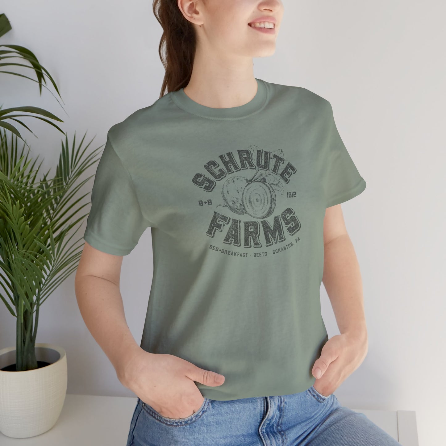 Person wearing a Sage green t-shirt featuring a Schrute Farms logo from the TV show The Office designed by ScriptedBrand in a vintage distressed style.