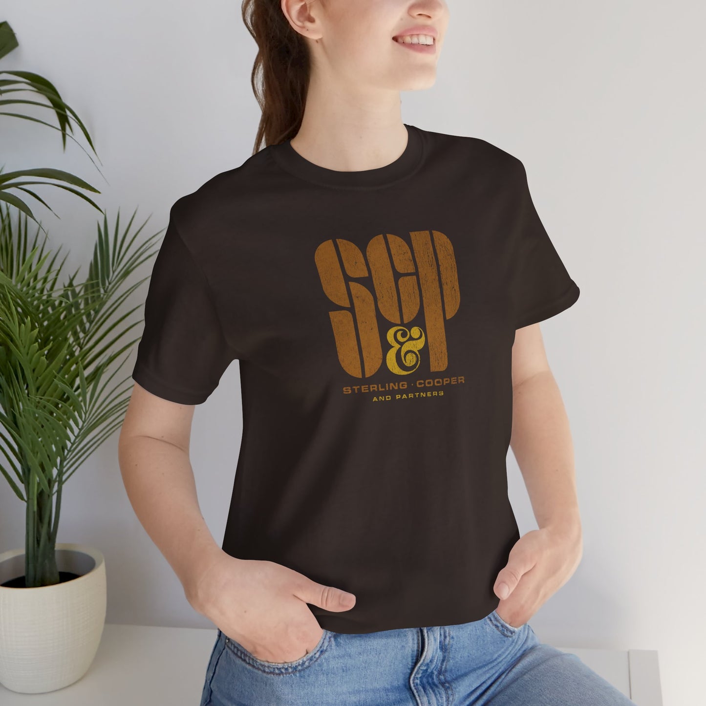 Female model wearing a brown colored soft unisex Bella+Canvas 3001 short-sleeve t-shirt featuring the Sterling Cooper & Partners logo graphic from the TV Show Mad Men in a vintage distressed style, sold by ScriptedBrand.