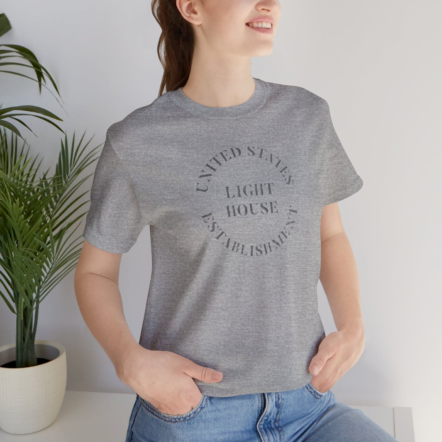 Person wearing a Heather Gray t-shirt featuring the United States Light House Establishment logo graphic worn by Robert Pattinson in the movie The Lighthouse in a vintage distressed style.