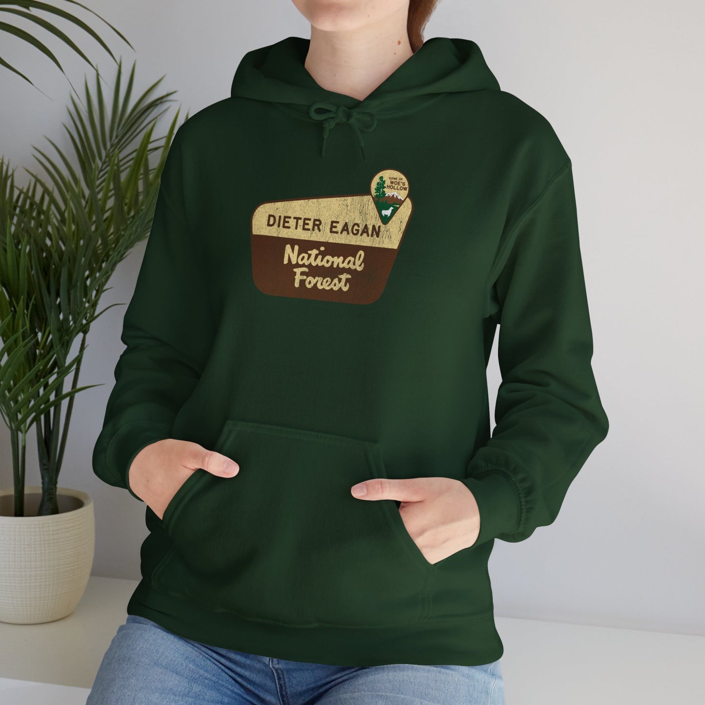 Female model wearing a Forest green colored Soft Unisex Hoodie Heavy Blend Hooded Sweatshirt featuring the logo graphic of the Dieter Eagan National Forest inspired by the TV show Severance in a vintage distressed ink-stamped worn-in style.