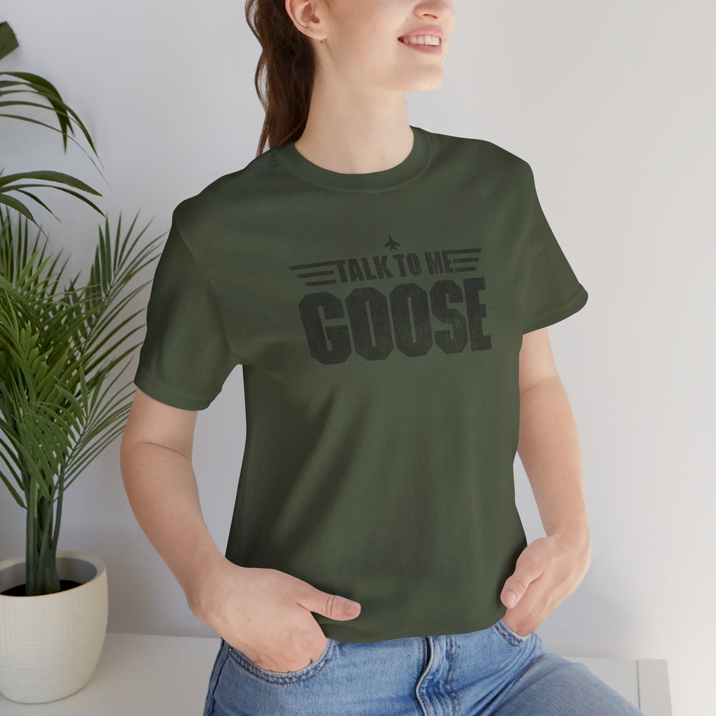 Female model wearing a military green soft unisex Bella+Canvas 3001 short-sleeve t-shirt featuring a graphic designed by ScriptedBrand in a vintage distressed style with the text TALK TO ME GOOSE, inspired by the Movie Top Gun.